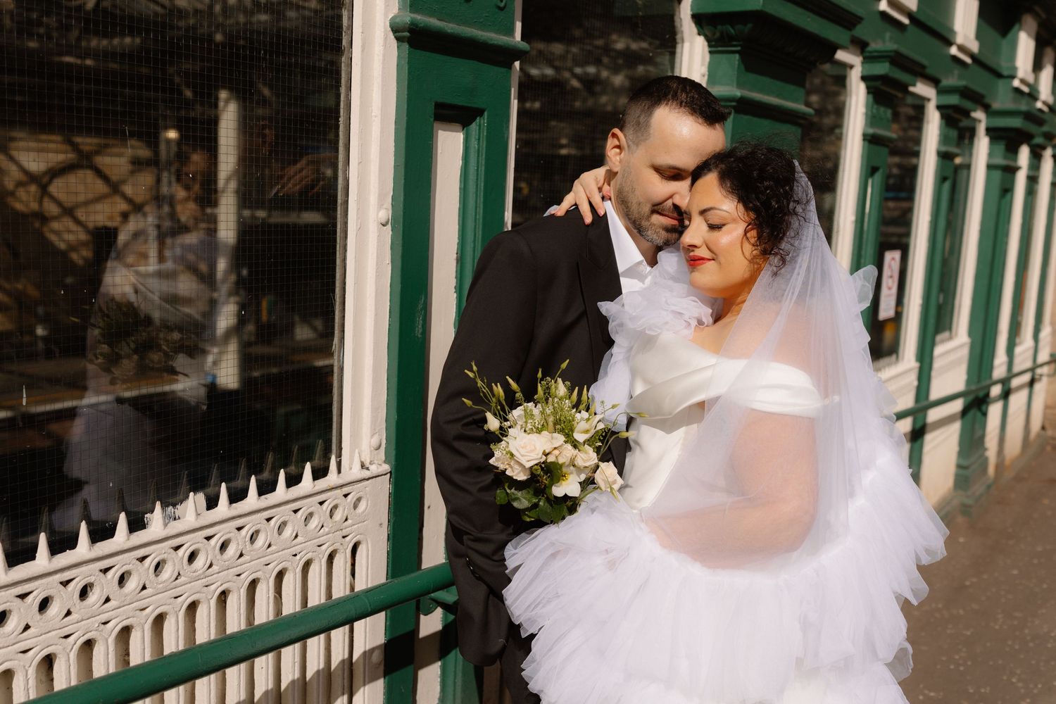 Bride in white gown holding bouquet embraces groom in black suit outdoors near green and white railing.