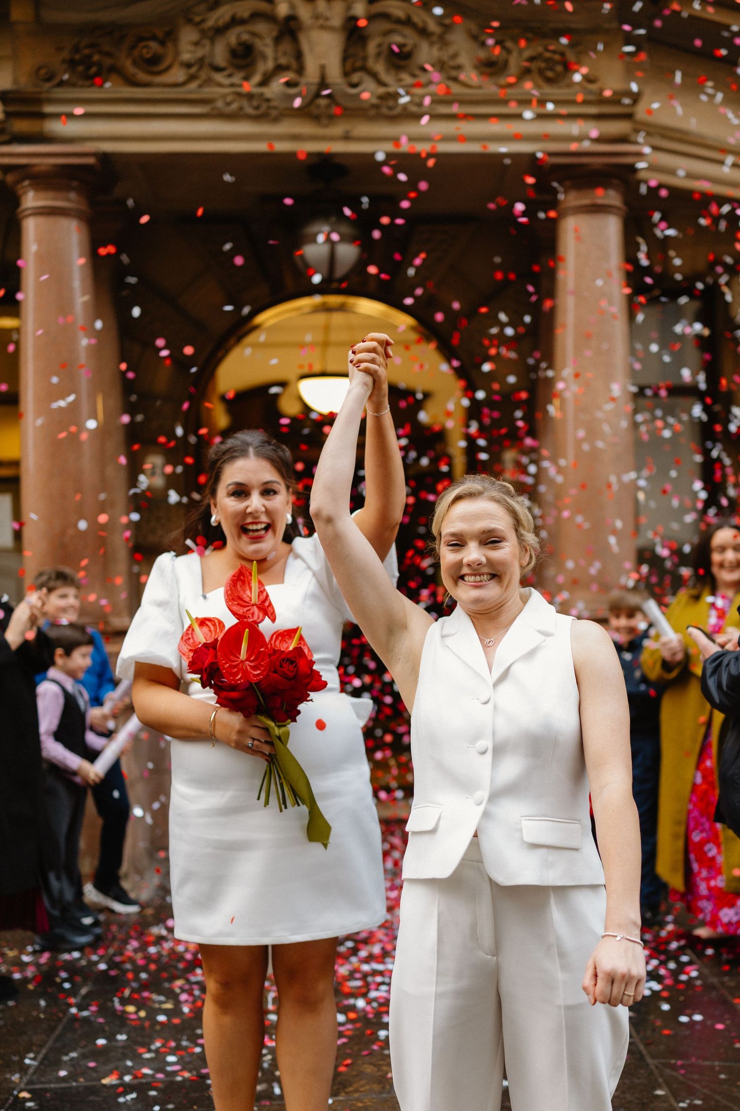 Two brides in white celebrate their wedding with raised hands and red bouquet amid colorful confetti outside a grand building.