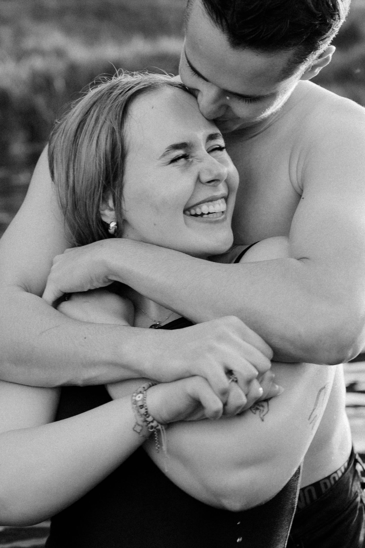 Black and white photo of a happy couple embracing outdoors, woman laughing joyfully as man hugs her from behind.