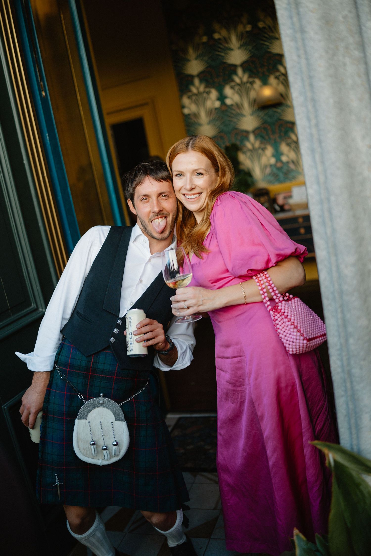 Man in tartan kilt and woman in pink dress smile together at an outdoor wedding event.
