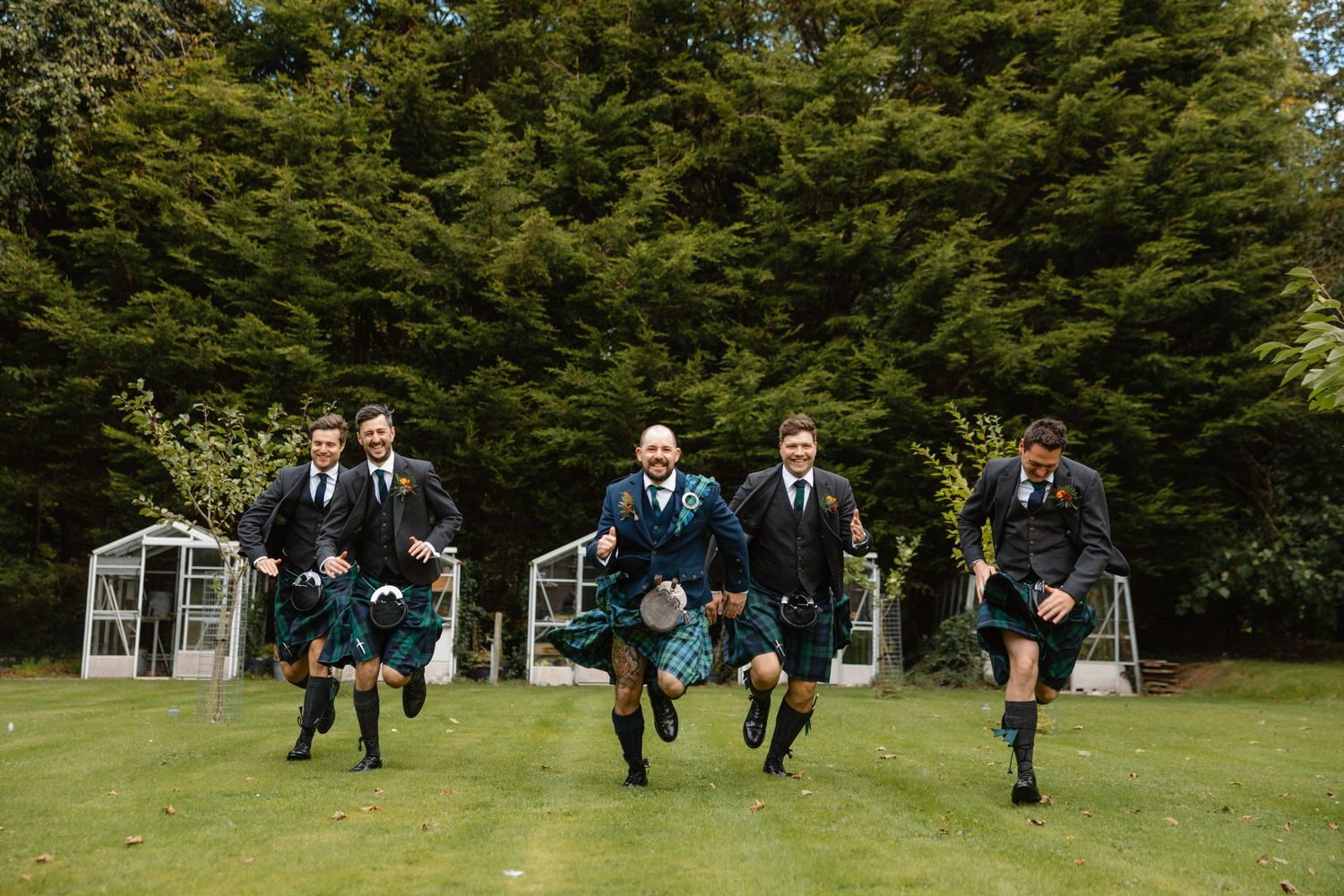 Groomsmen in kilts running across a green lawn with tall trees in the background at a Scottish wedding.