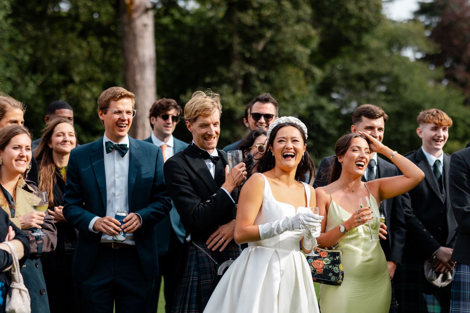 Laughing bride in white dress with guests in formal attire enjoying outdoor wedding reception drinks.