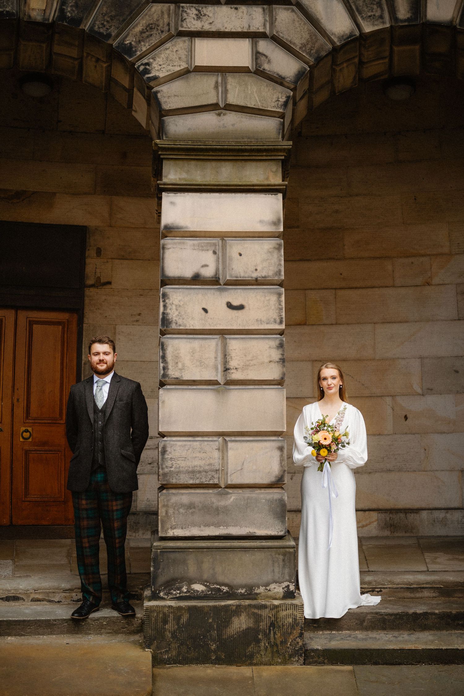 Bride and groom stand on either side of a stone column at a historic building entrance.