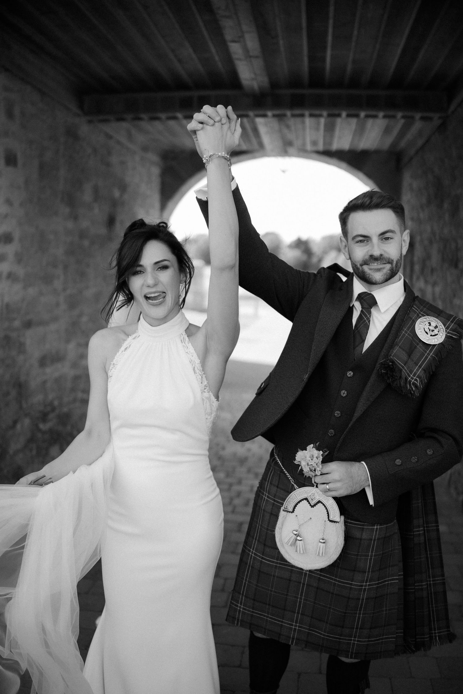 Black and white photo of a bride and groom in Scottish kilt raising hands in celebration at wedding.