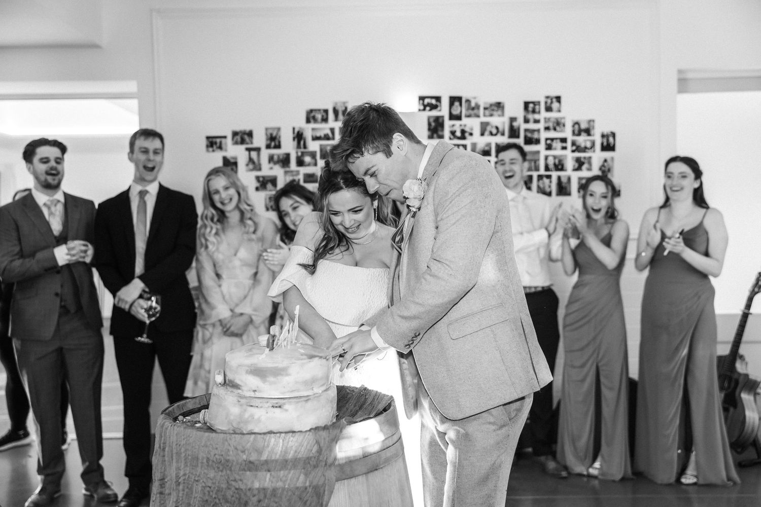 Bride and groom cutting wedding cake surrounded by smiling wedding party guests indoors.