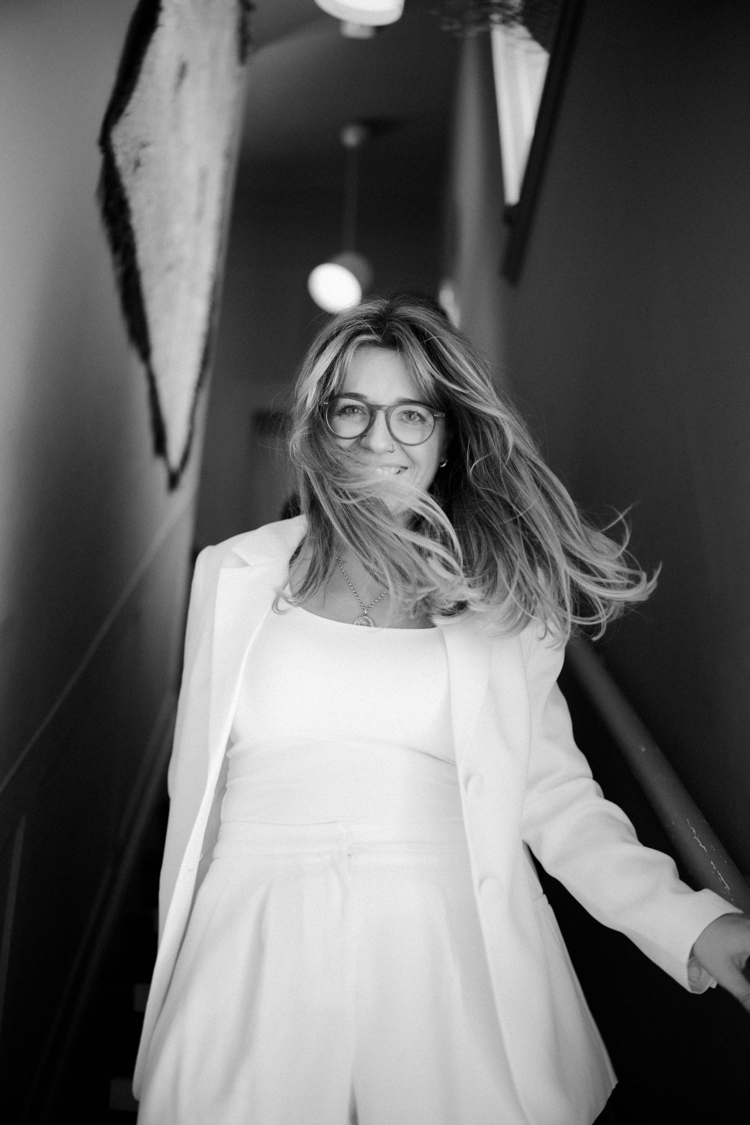 Black and white photo of a smiling woman with flowing hair wearing white blazer and glasses in a hallway.
