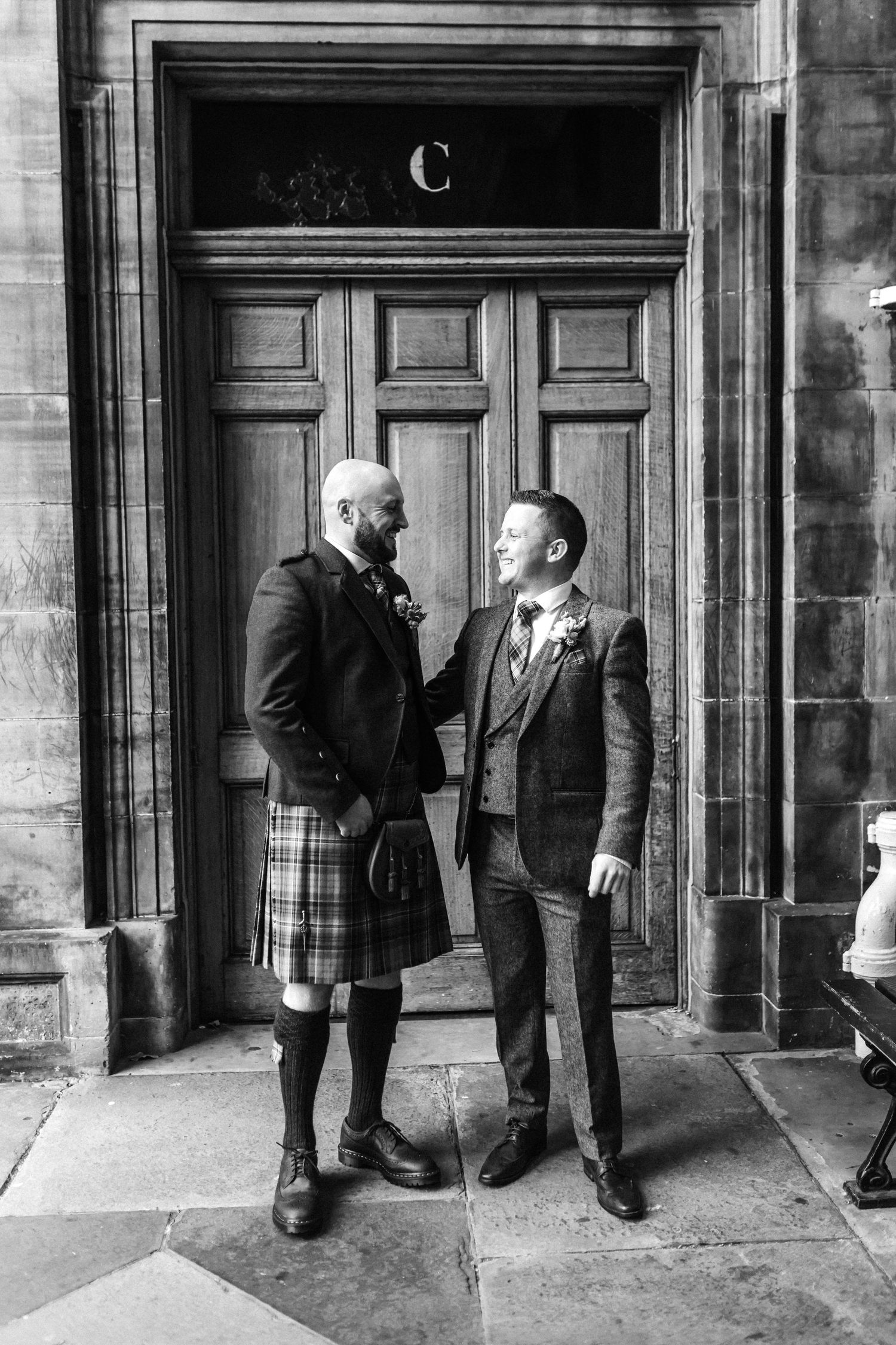 Groom in tartan kilt and suited best man share a moment outside ornate stone building in black and white photo.