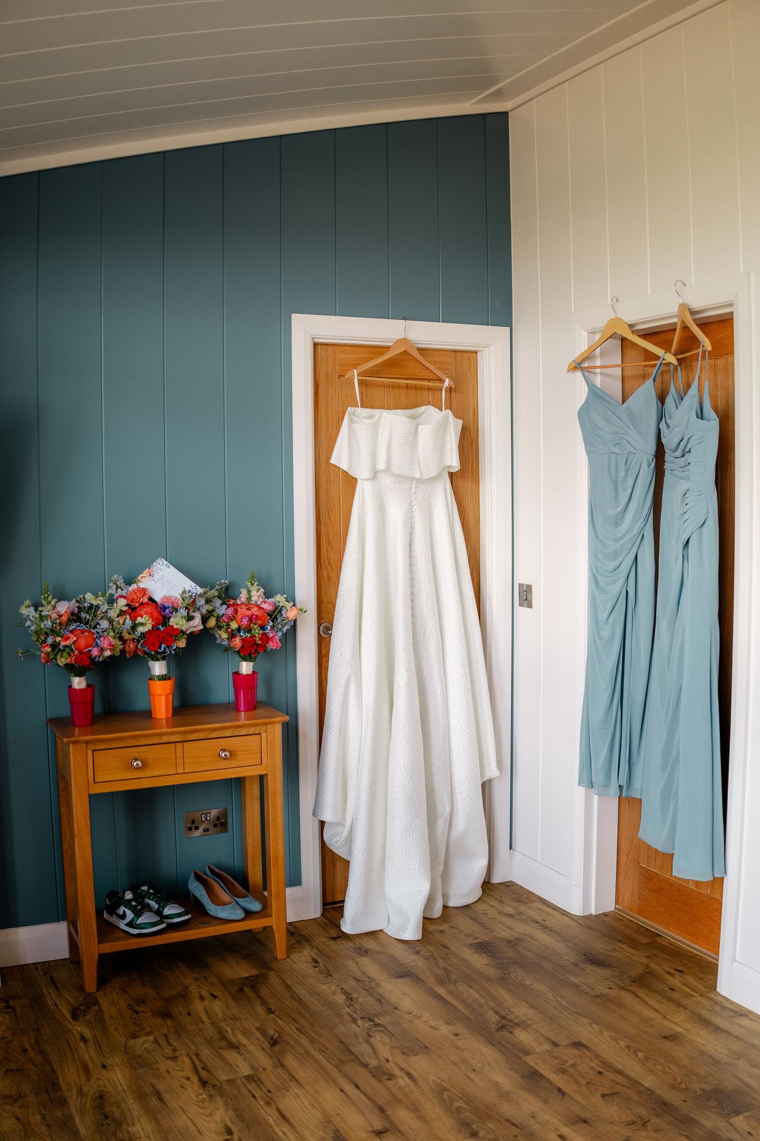 Wedding attire displayed on hangers against a teal wall, including a white dress and blue bridesmaid dresses.