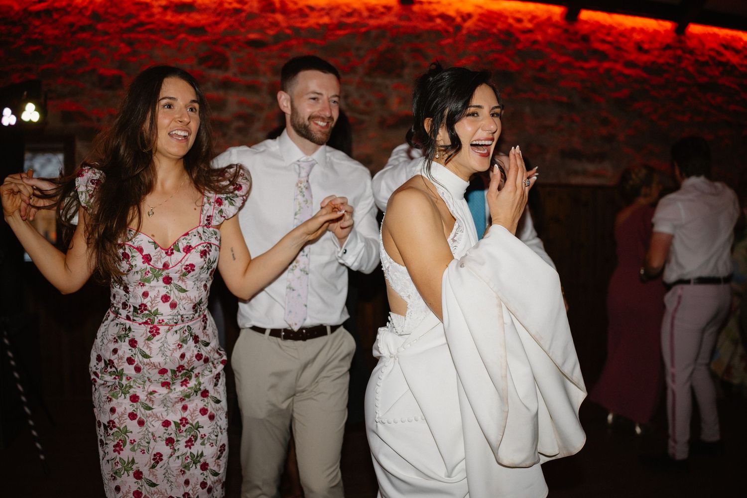 Wedding party dancing and celebrating under red mood lighting in a rustic venue.