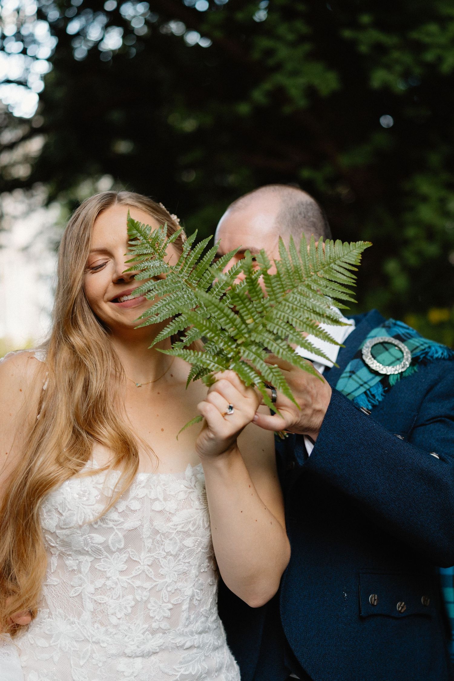 Playful wedding couple poses with green fern leaves in an outdoor garden setting.