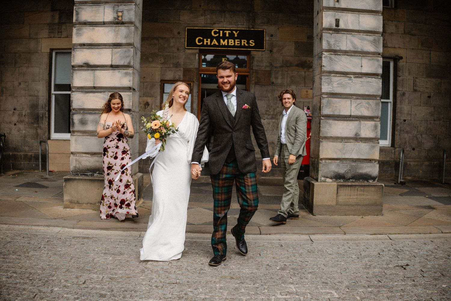 Newlywed couple holding hands and smiling outside Edinburgh City Chambers with wedding guests behind.