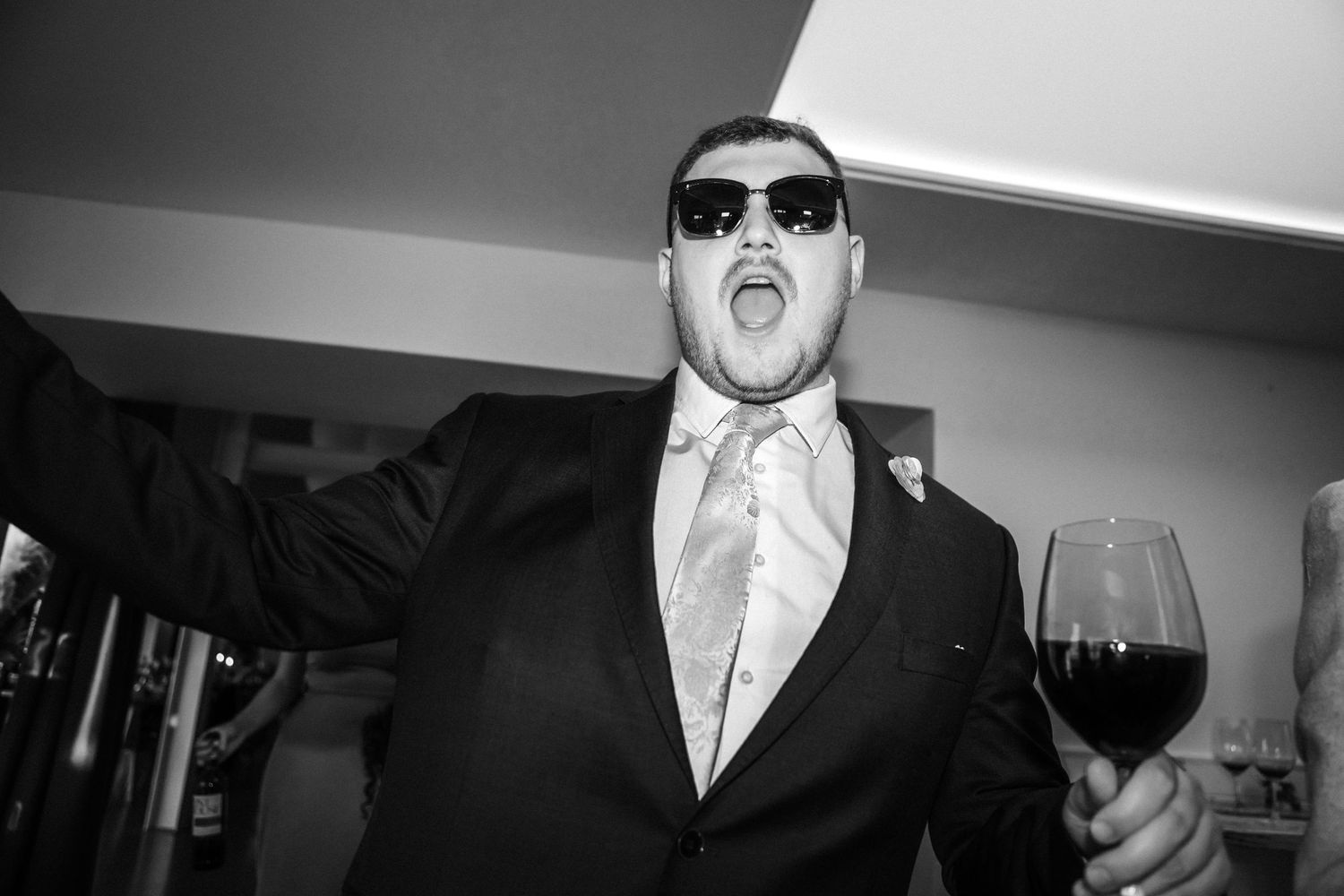 Man in suit and sunglasses holding red wine glass at party, mouth open in celebration, black and white photo.