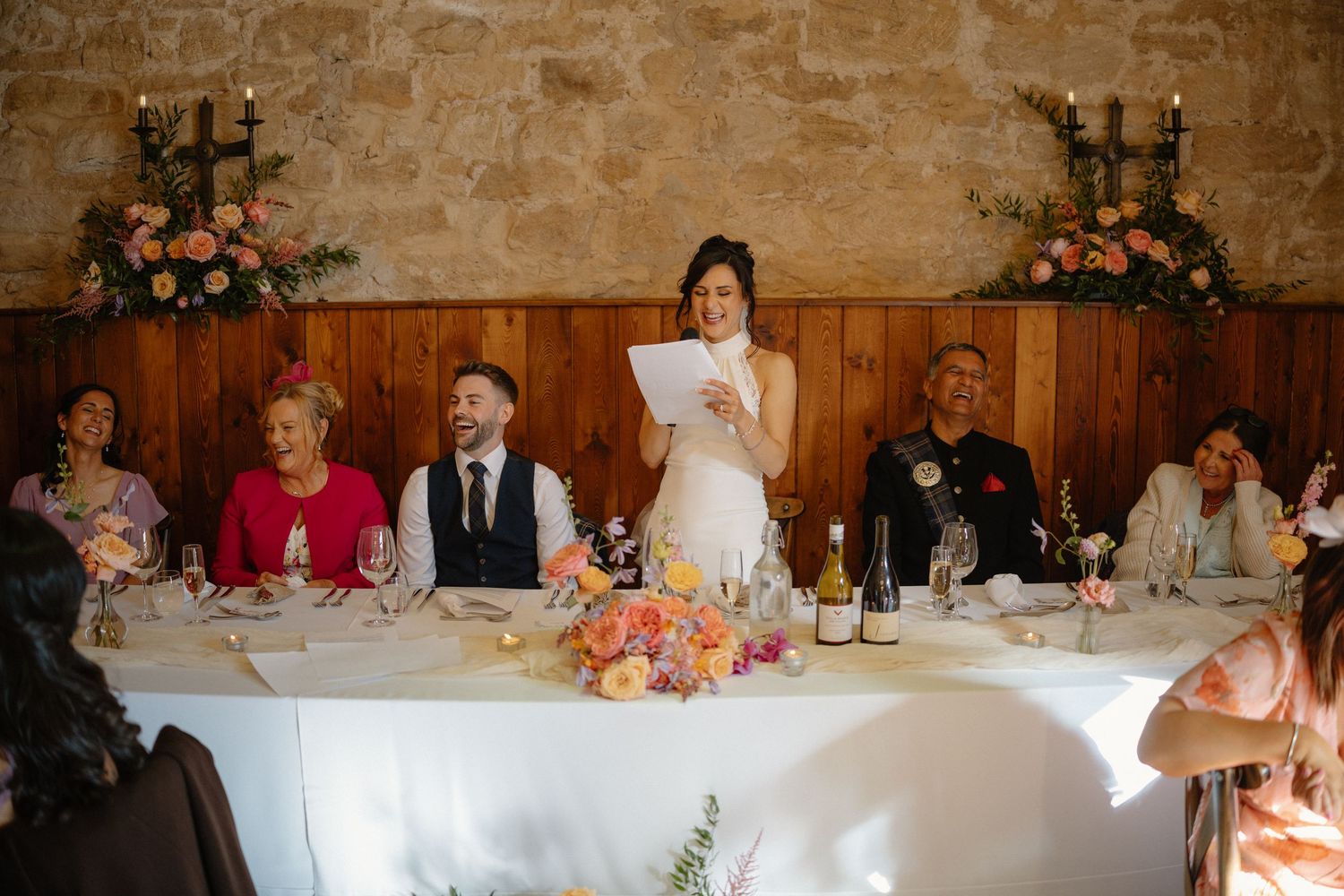 Bride in white gown giving a speech at the wedding head table decorated with colorful floral arrangements.