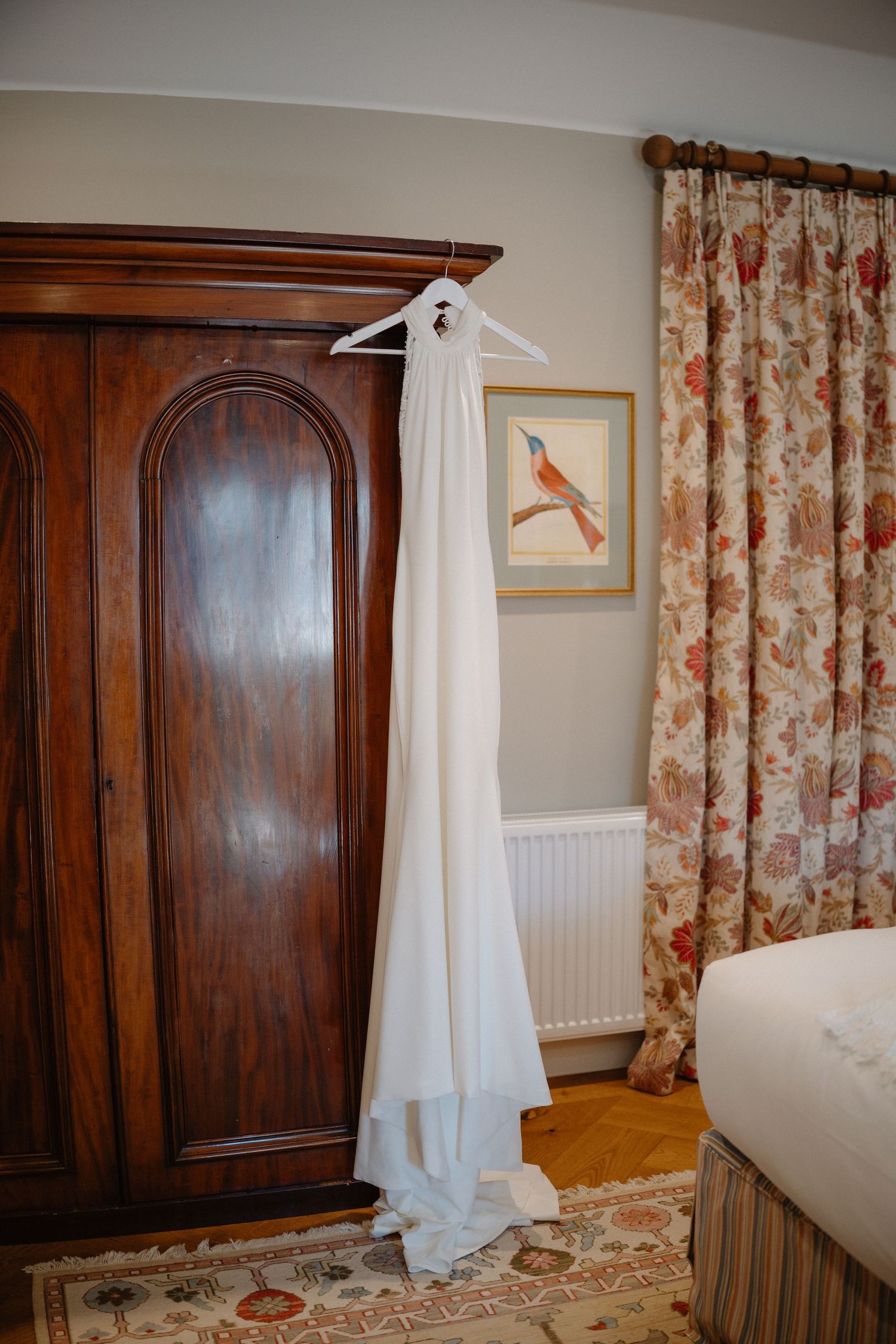 White wedding dress hanging on a wooden wardrobe in an elegant bedroom with floral curtains.