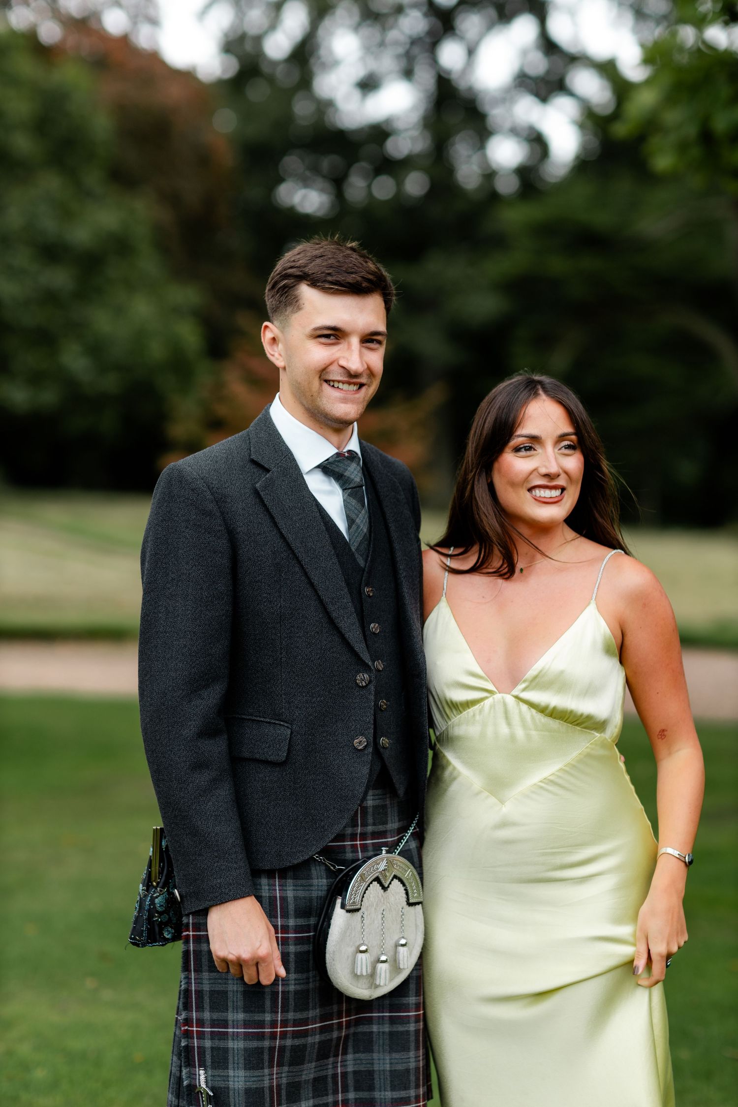 Smiling couple at Scottish wedding, man in traditional tartan kilt and jacket, woman in elegant sage green satin slip dress.