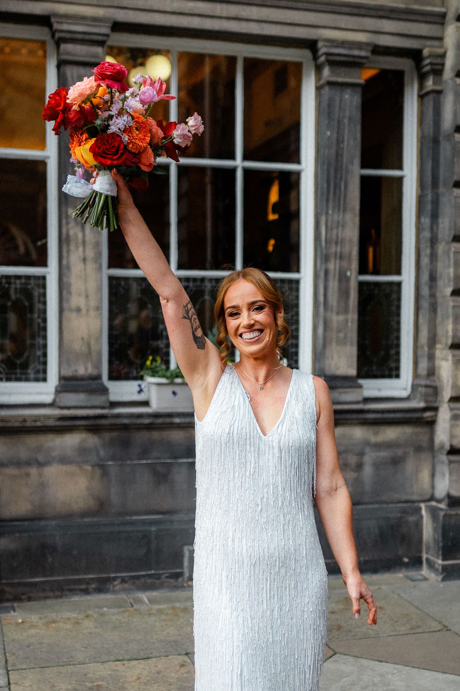 Joyful bride in silver fringe dress raising colorful bouquet of red and orange flowers outdoors.