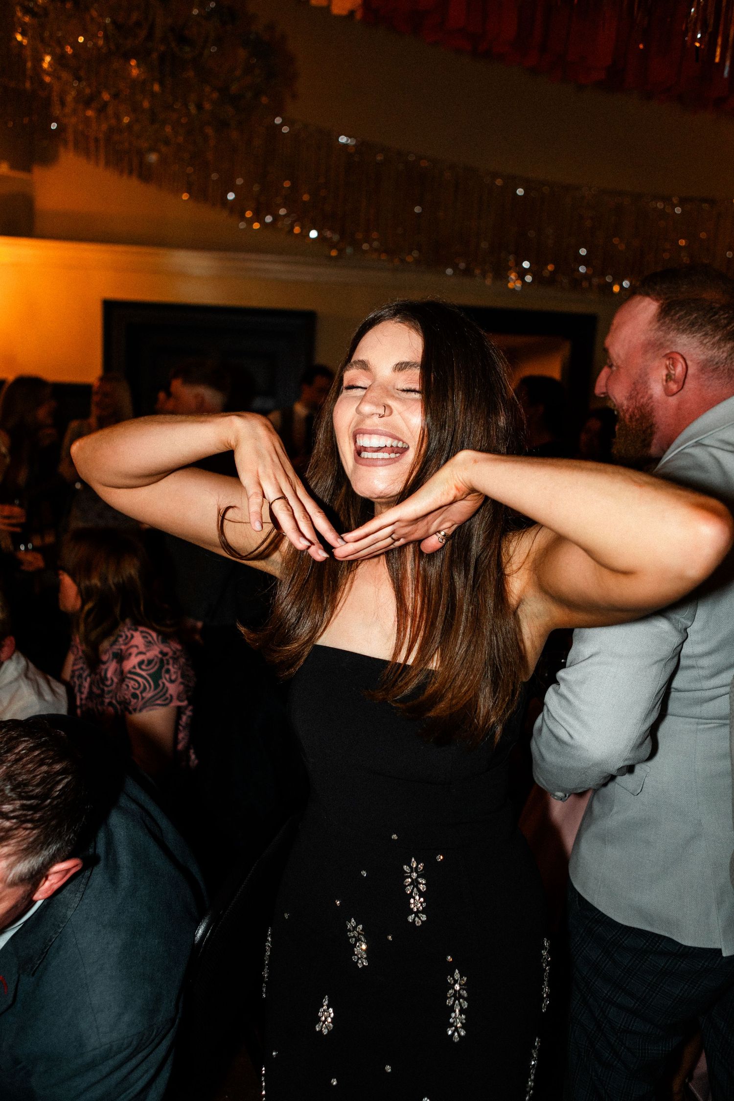 Joyful woman in black dress laughing and dancing at lively wedding reception party under warm golden lights.