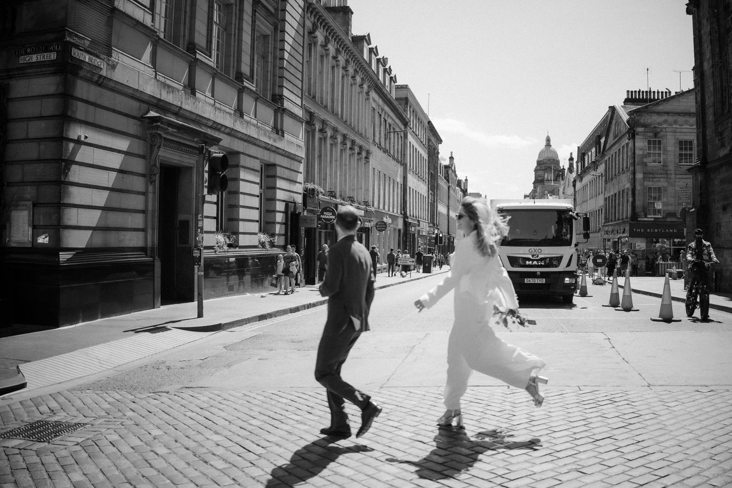 Black and white photo of bride and groom crossing cobblestone street in city after wedding ceremony.