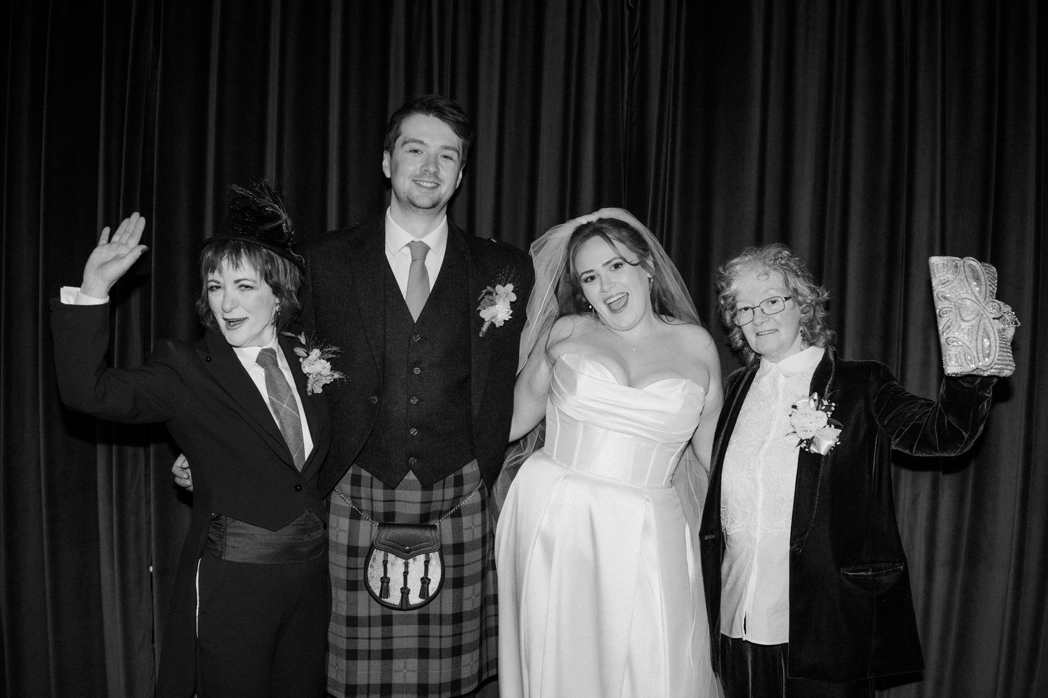 Black and white photo of bride, groom in kilt, and two guests celebrating joyfully at wedding reception.