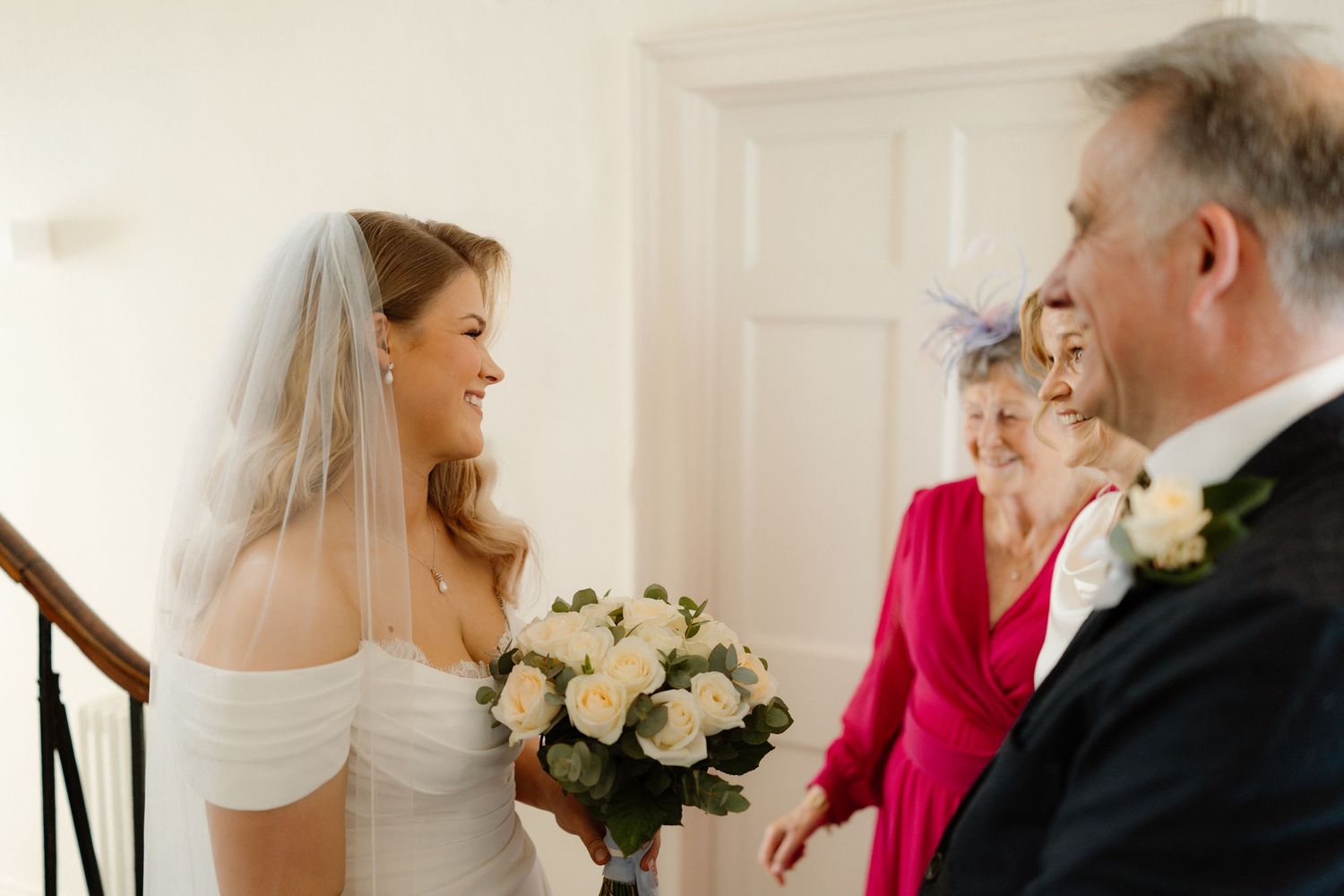 Smiling bride in white off-shoulder gown and veil holds white rose bouquet greeting family before wedding ceremony.