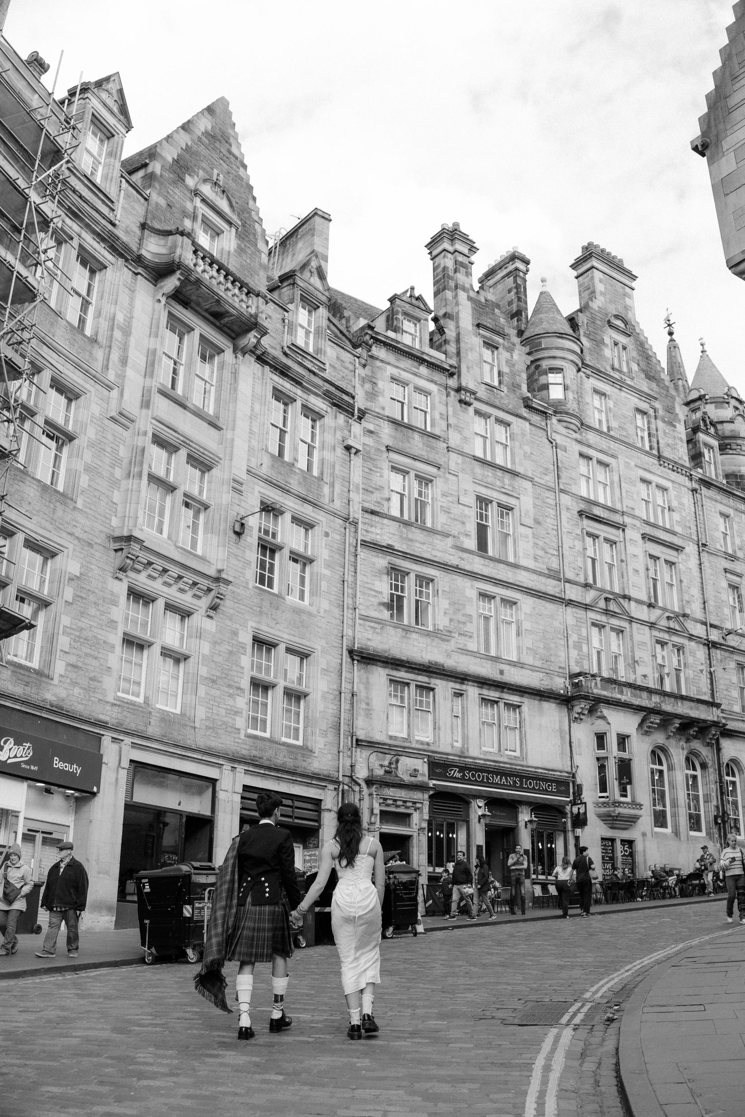 Black and white photo of couple in traditional Scottish kilts walking on Royal Mile, Edinburgh's historic cobblestone street.