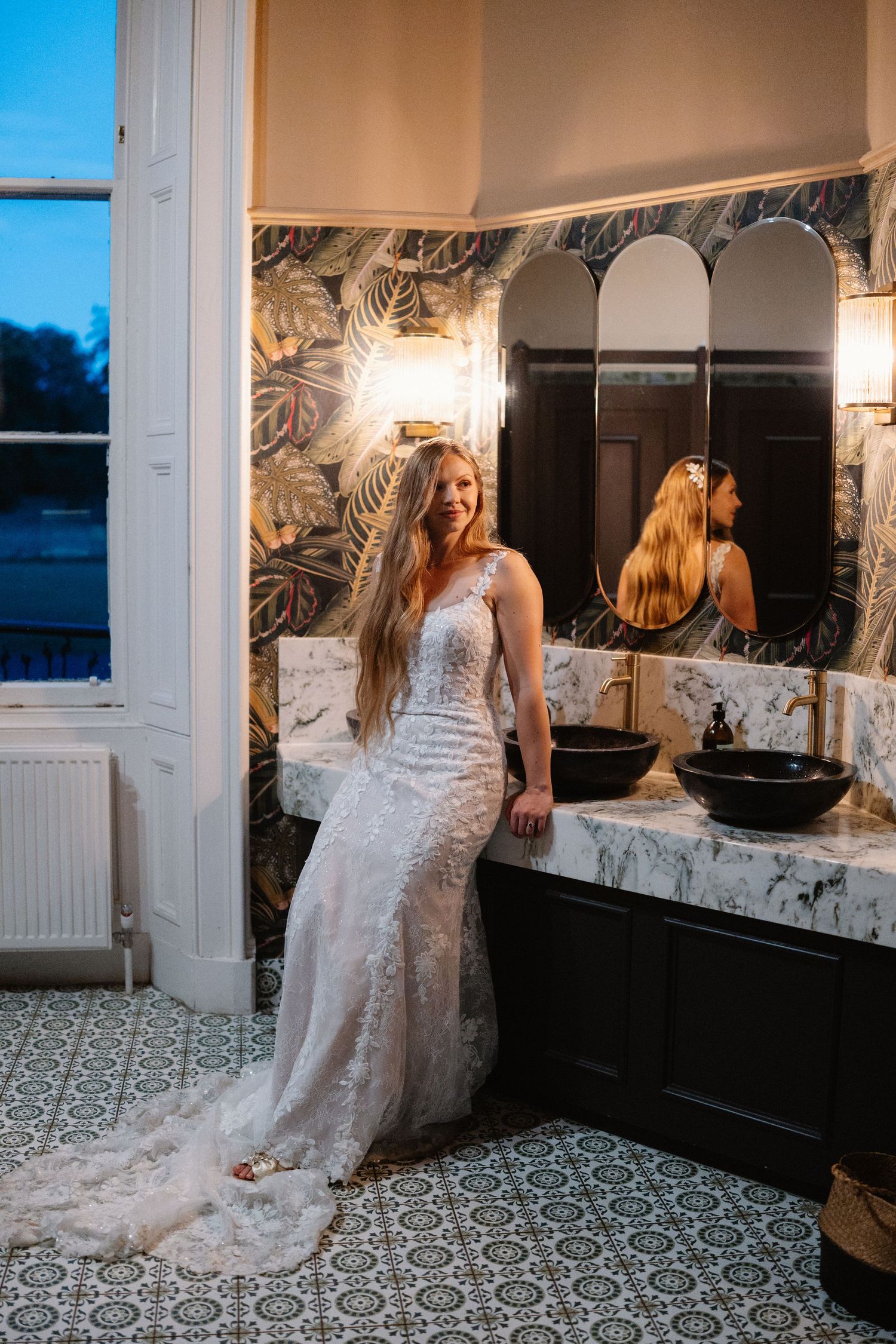 Bride in lace wedding gown posing in stylish bathroom with marble countertop and tropical wallpaper.