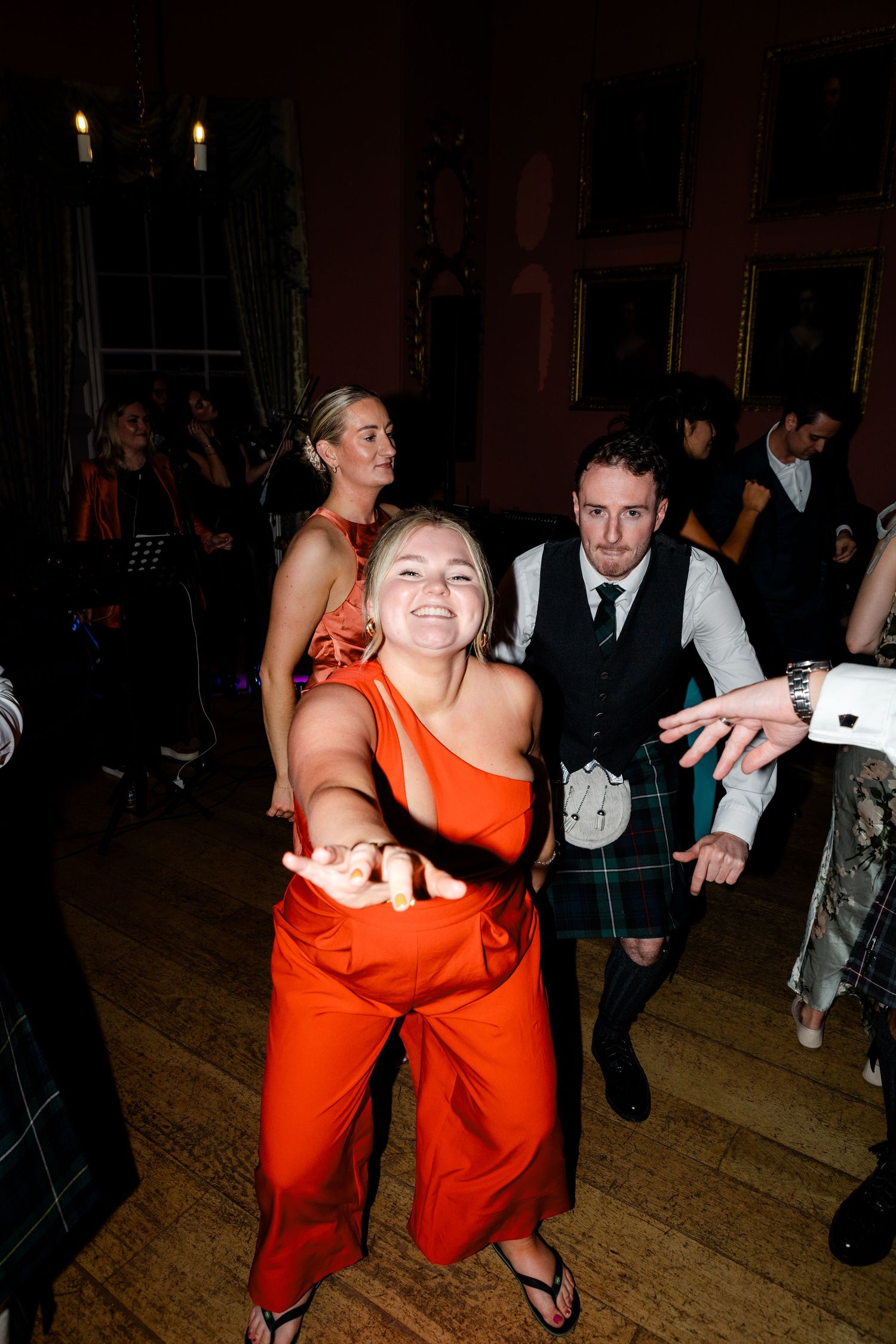Wedding guests in formal attire dance energetically, with a woman in an orange jumpsuit leading the fun.