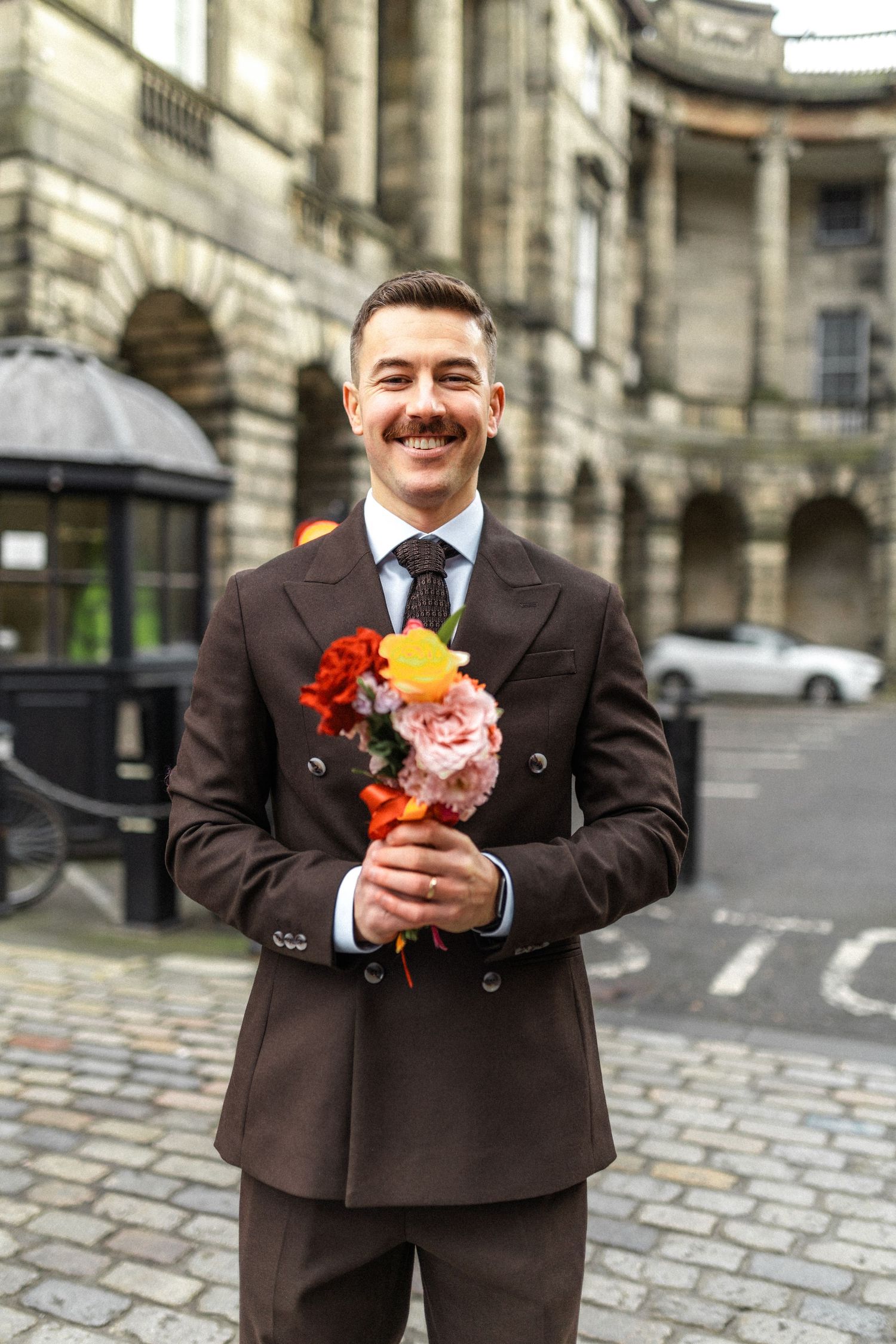 Smiling groom in brown double-breasted suit holding colorful bridal bouquet outside historic stone building.