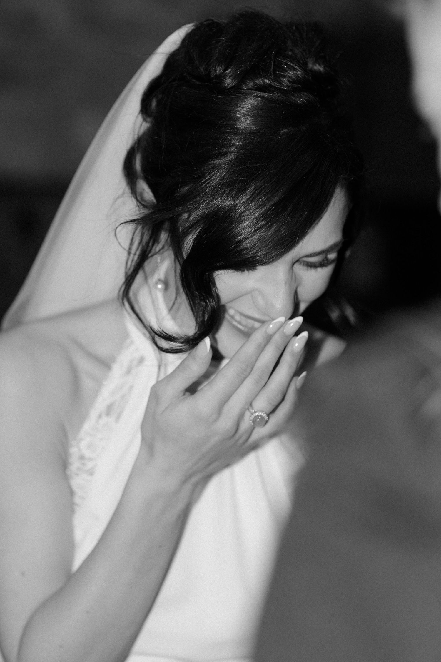 Emotional bride in white veil laughing with hand over mouth showing engagement ring in black and white photo.