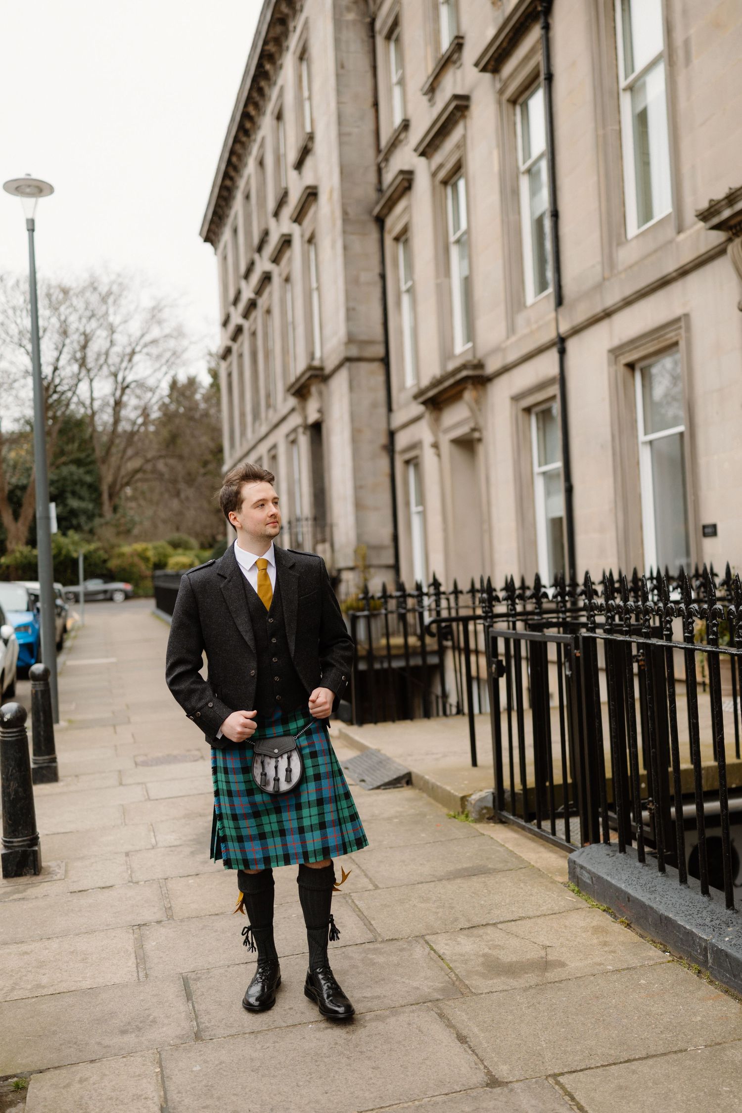 A young man in traditional Scottish attire with a green tartan kilt and black jacket walking on a city sidewalk.