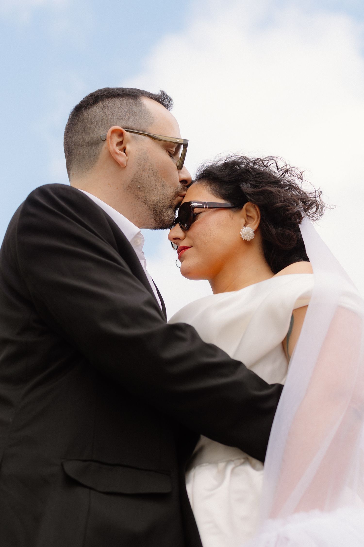 Groom in black suit kissing bride wearing sunglasses and white dress against a blue sky backdrop.