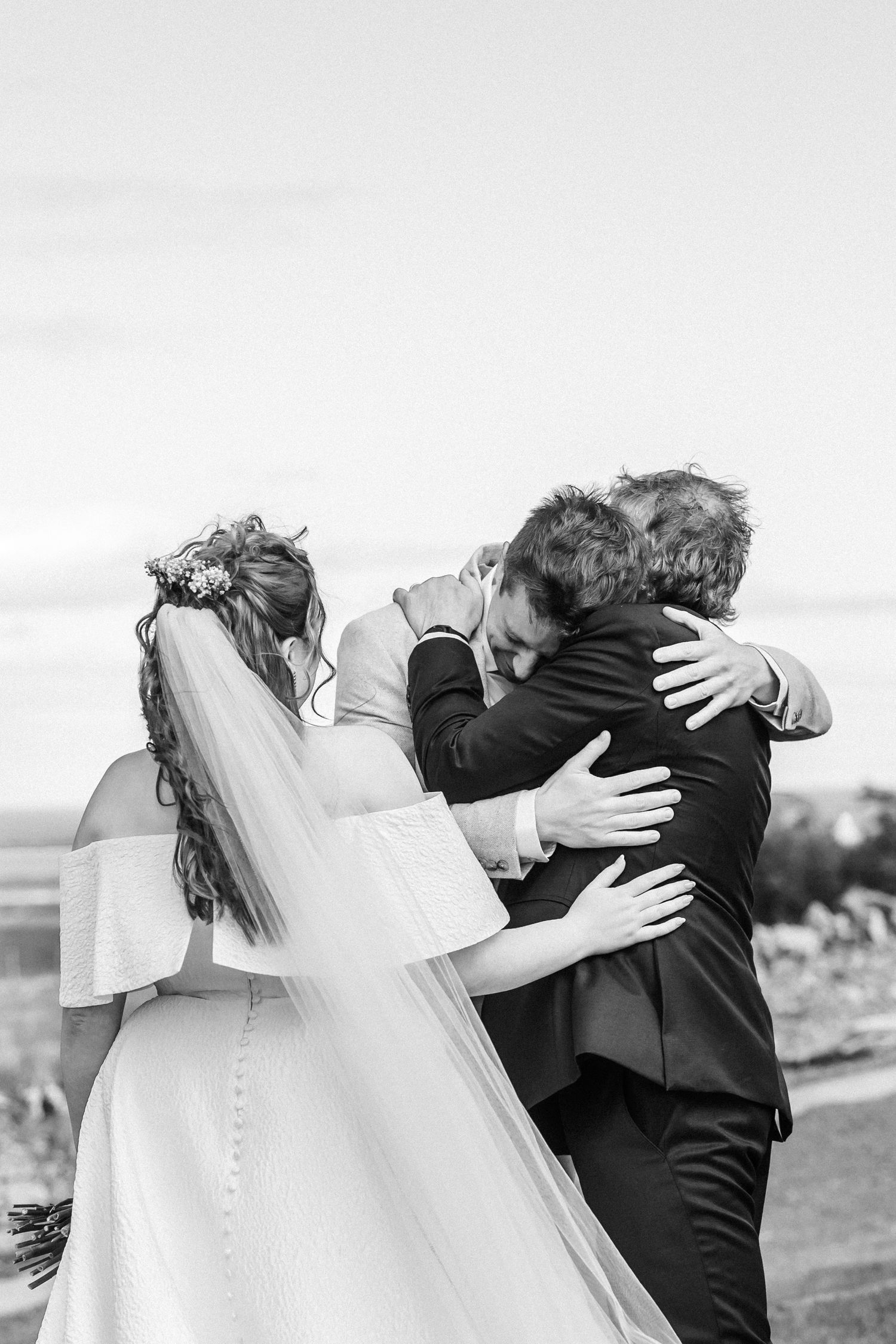 Black and white photo of bride and groom sharing emotional group hug at wedding ceremony.