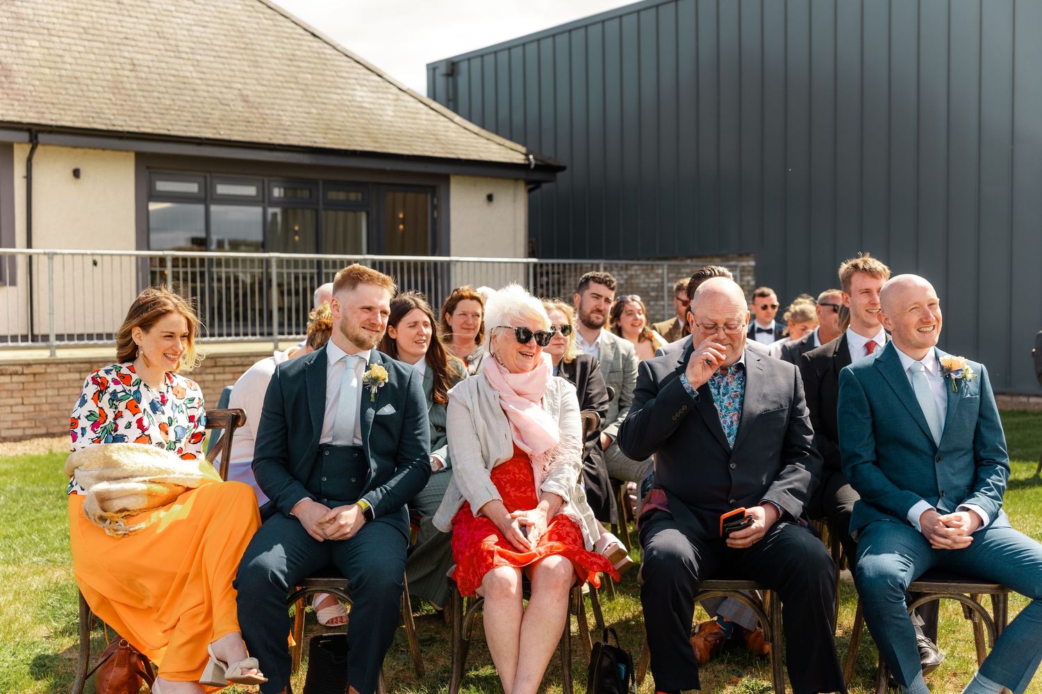 Wedding guests laugh and smile with emotion during an outdoor ceremony at a modern barn venue.