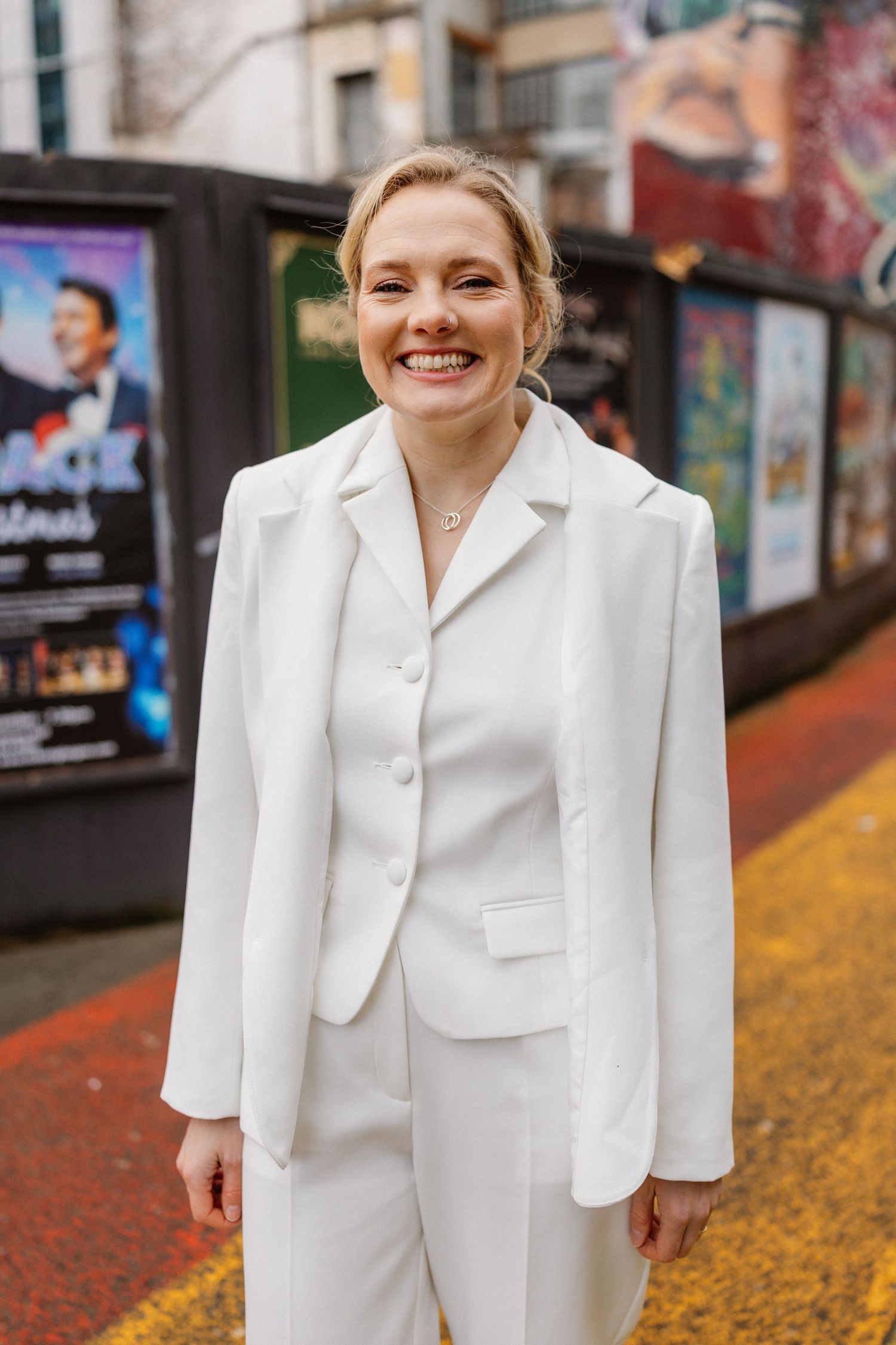 Smiling blonde bride in a white trouser suit standing on a colorful street with theater posters.
