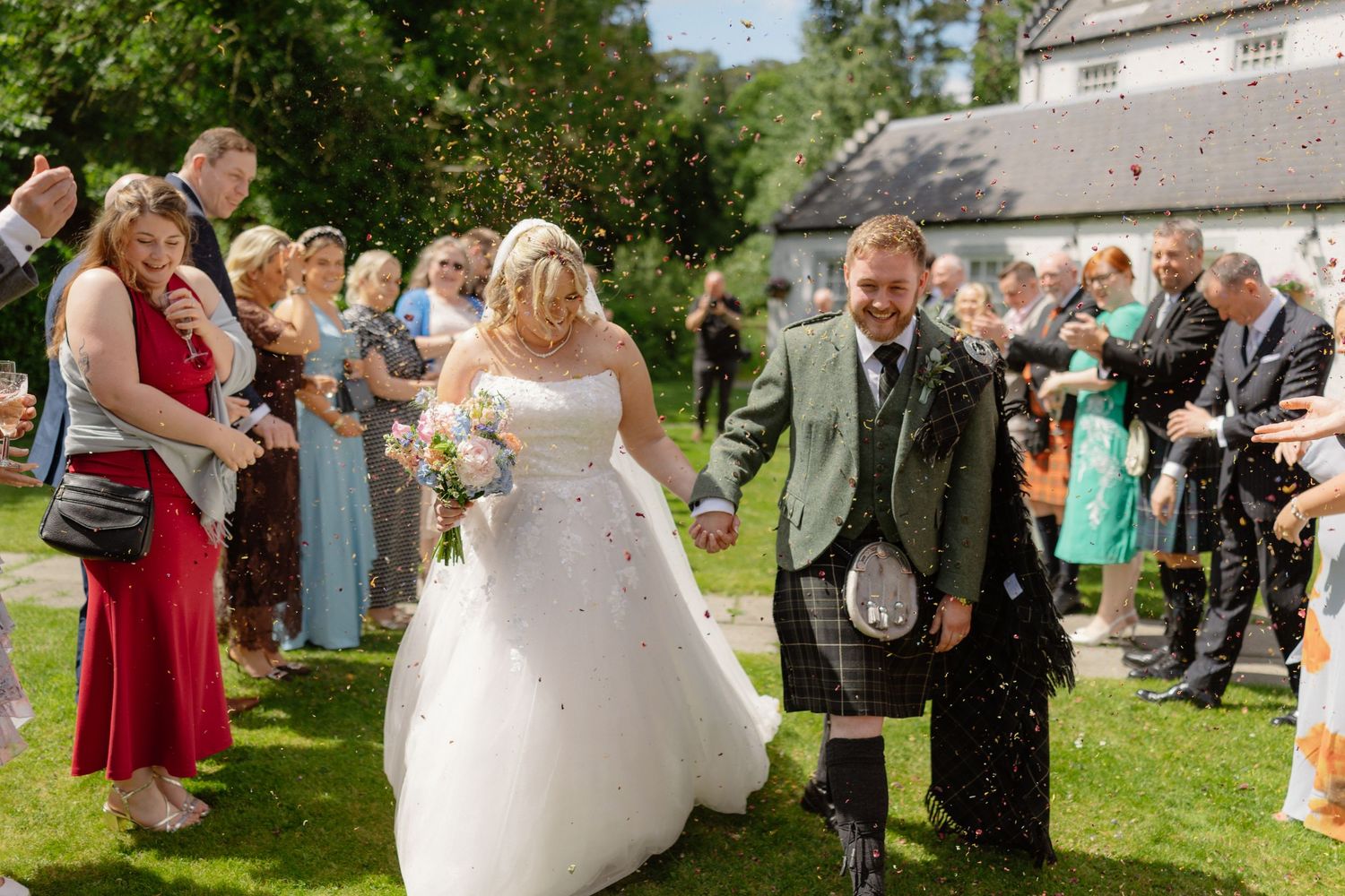 Bride and groom walk through confetti shower at outdoor Scottish wedding, groom wearing traditional tartan kilt.