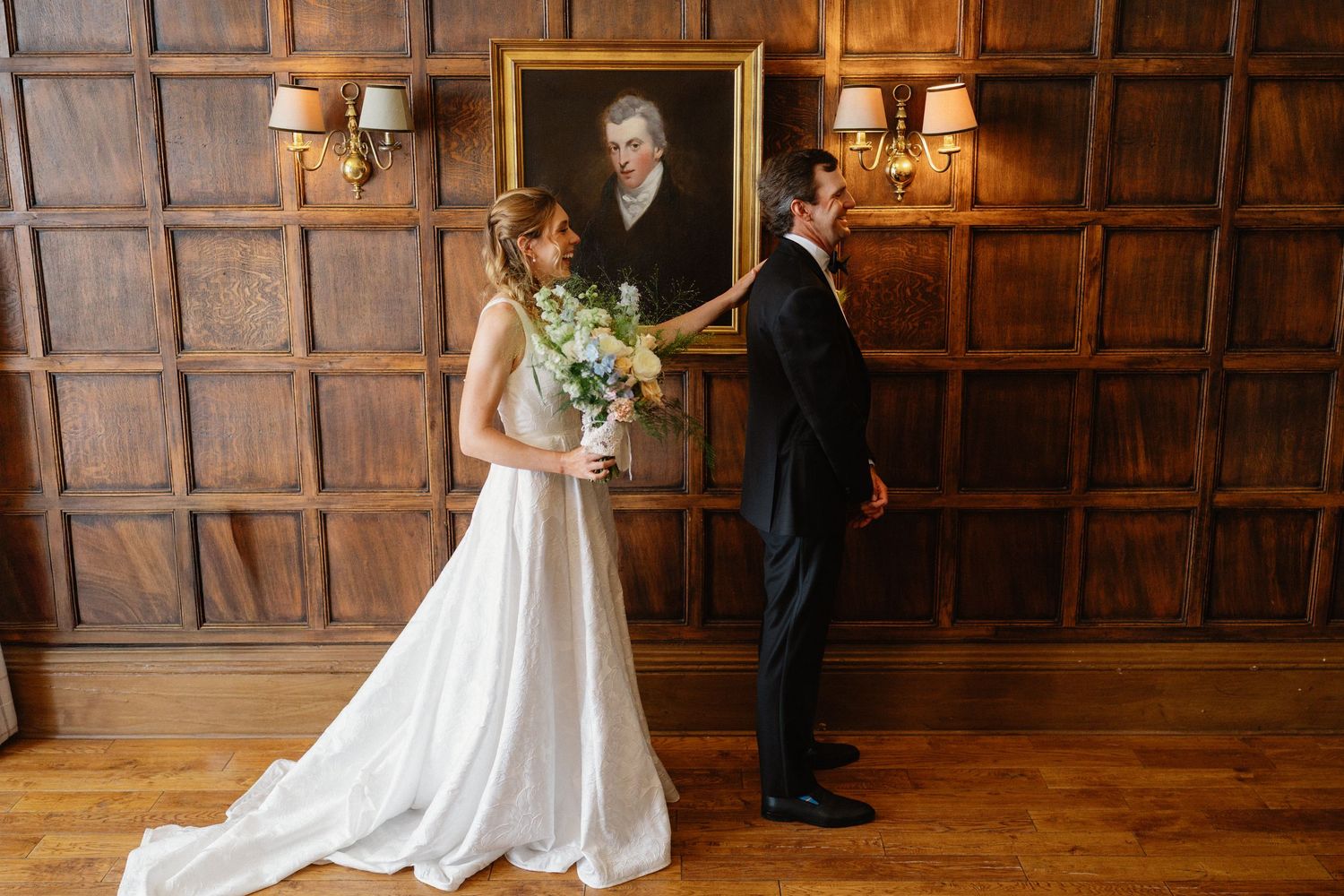 Bride and groom share a tender first look moment in an elegant wood-paneled room with a classic portrait painting.