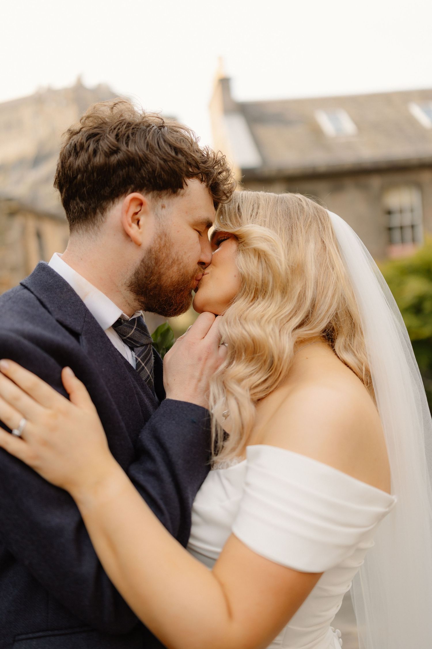 A bride and groom share a tender outdoor kiss, she in a white off-shoulder gown with veil, he in a navy suit.