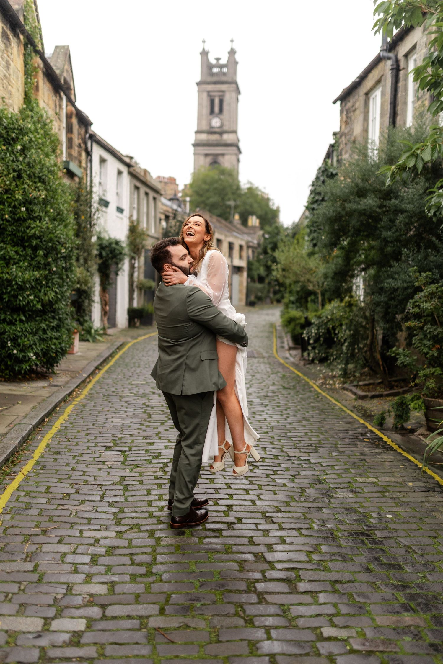 Romantic couple poses on a cobblestone street in Edinburgh with historic architecture in background.