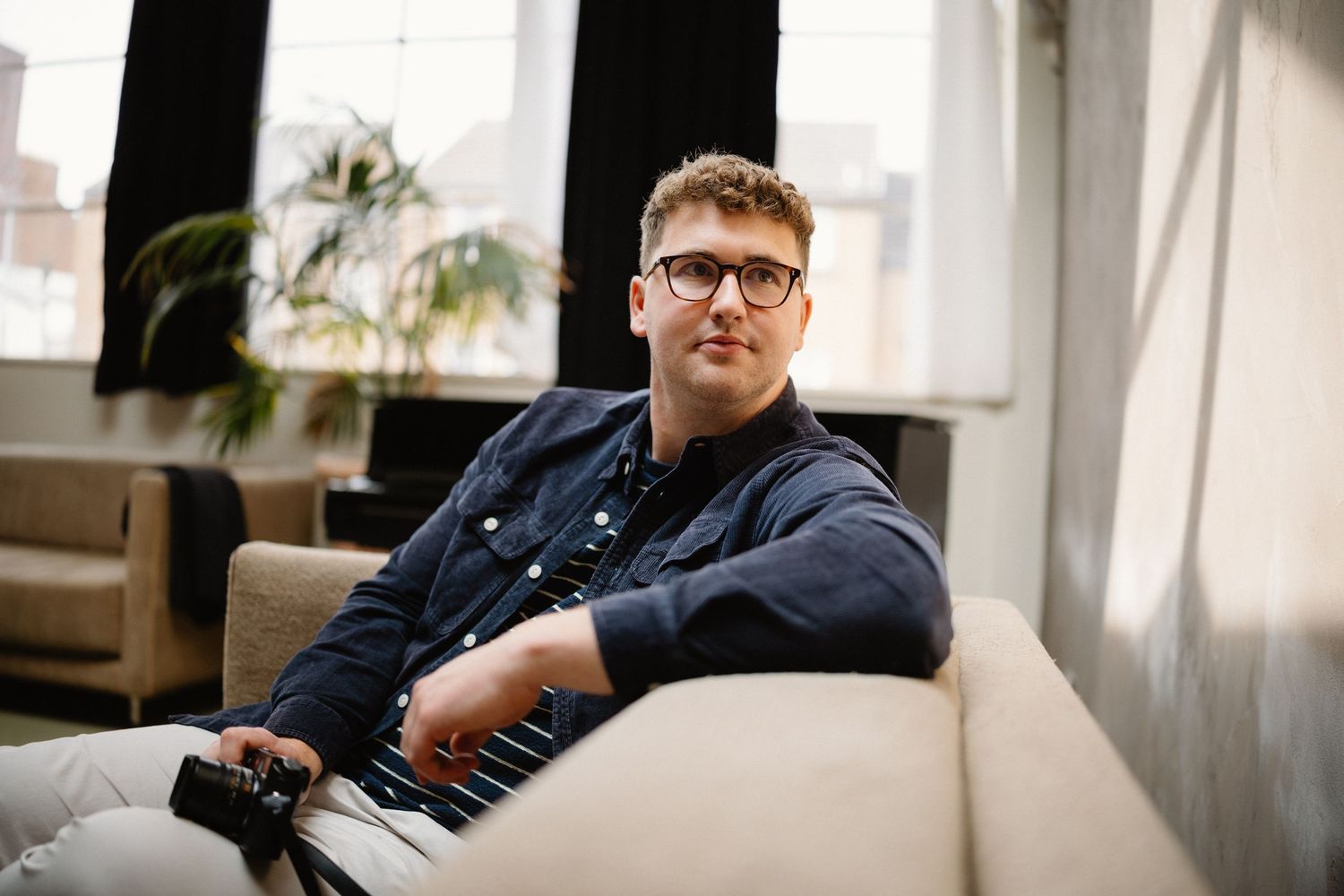 Person in denim jacket sitting casually on a couch in a bright room.