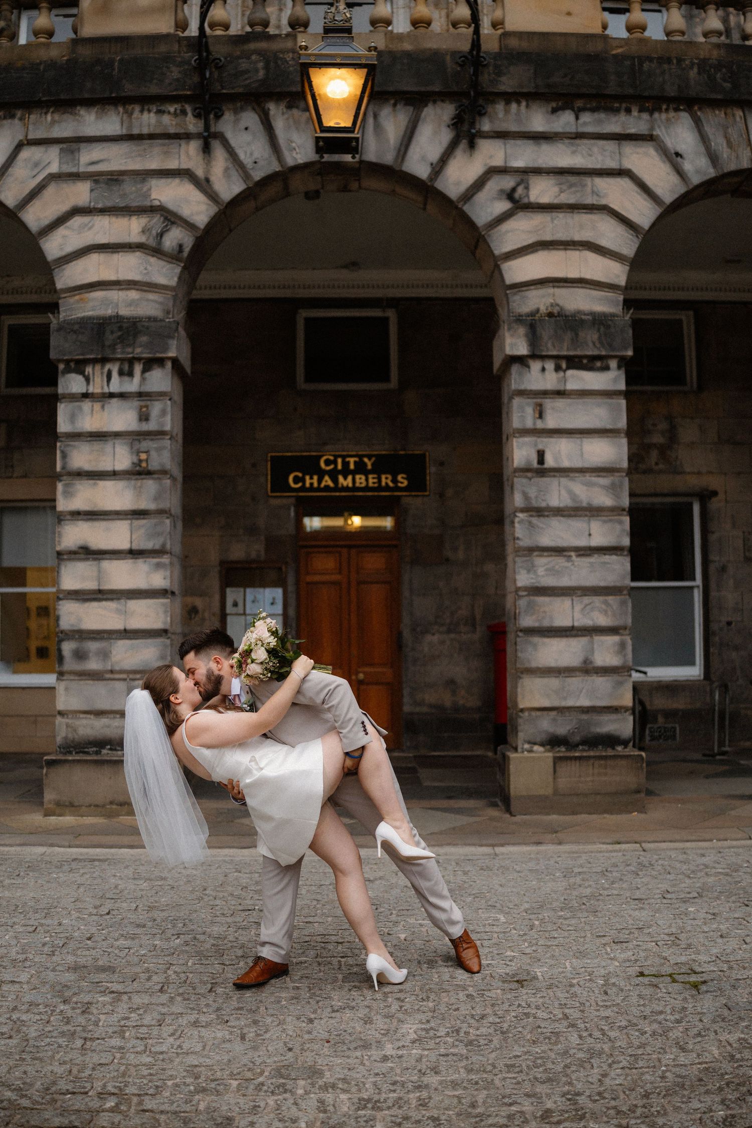 A couple in wedding attire playfully dances and dips outside a historic stone building with City Chambers signage.