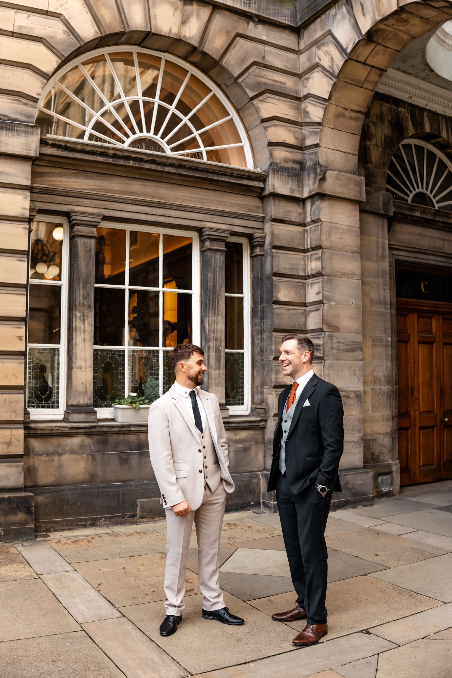 Two people in formal attire having a conversation outside a historic stone building with arched doorways.