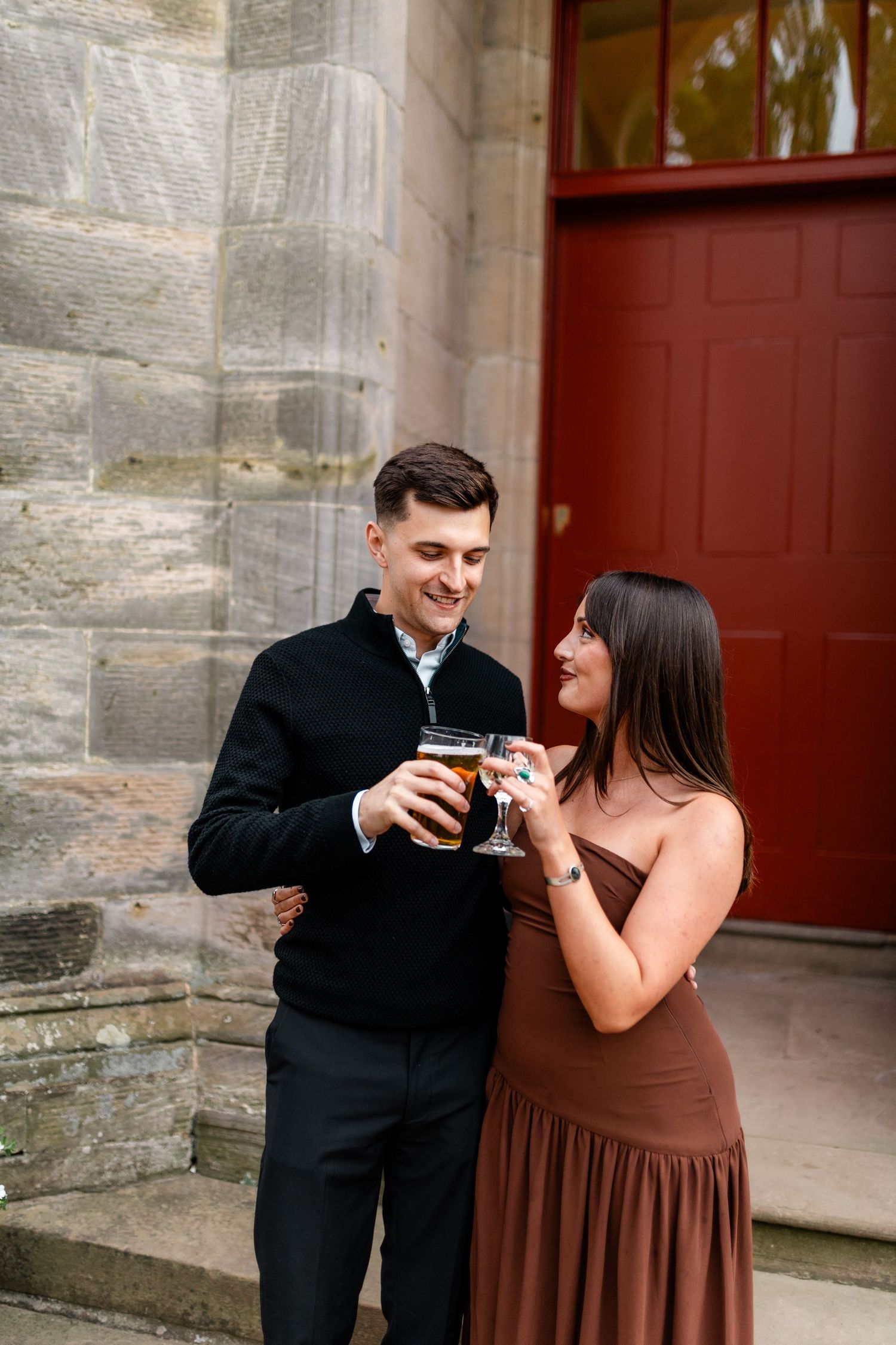 Couple shares drinks together in formal attire against a stone building exterior.