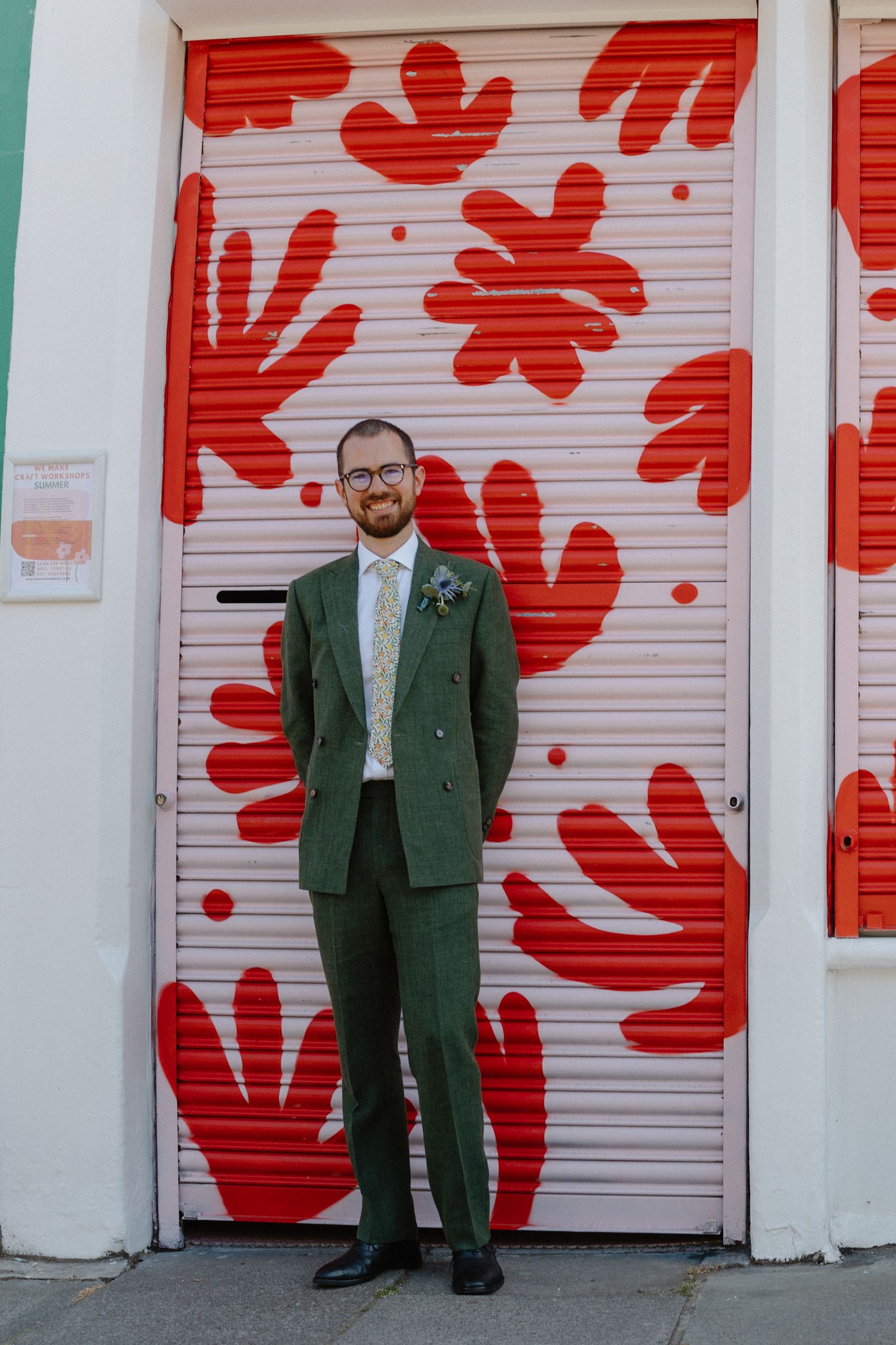 A person in a green suit stands against a wall featuring large red floral designs.