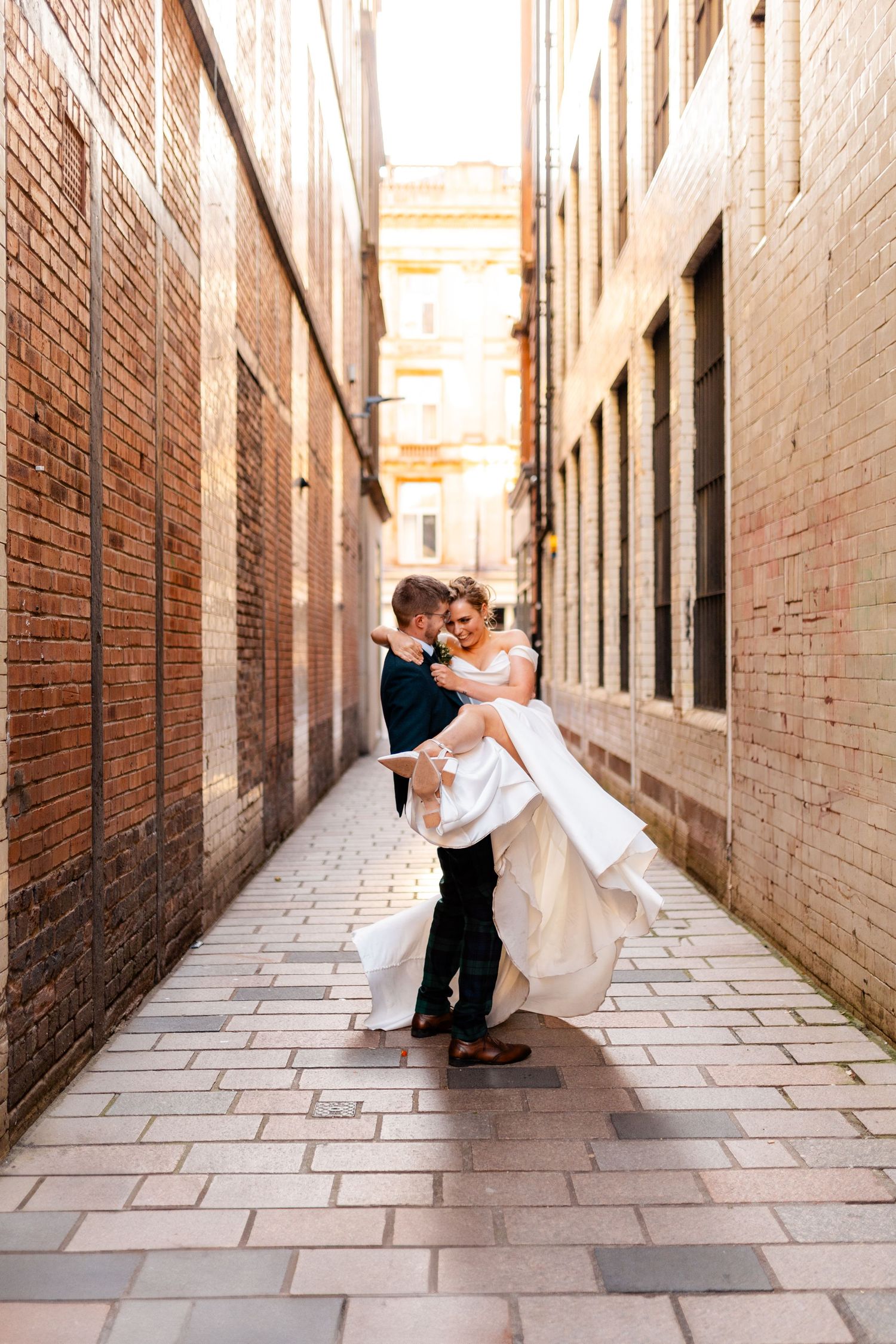 Groom lifting bride in white dress in a sunlit brick alley, sharing a joyful wedding moment.