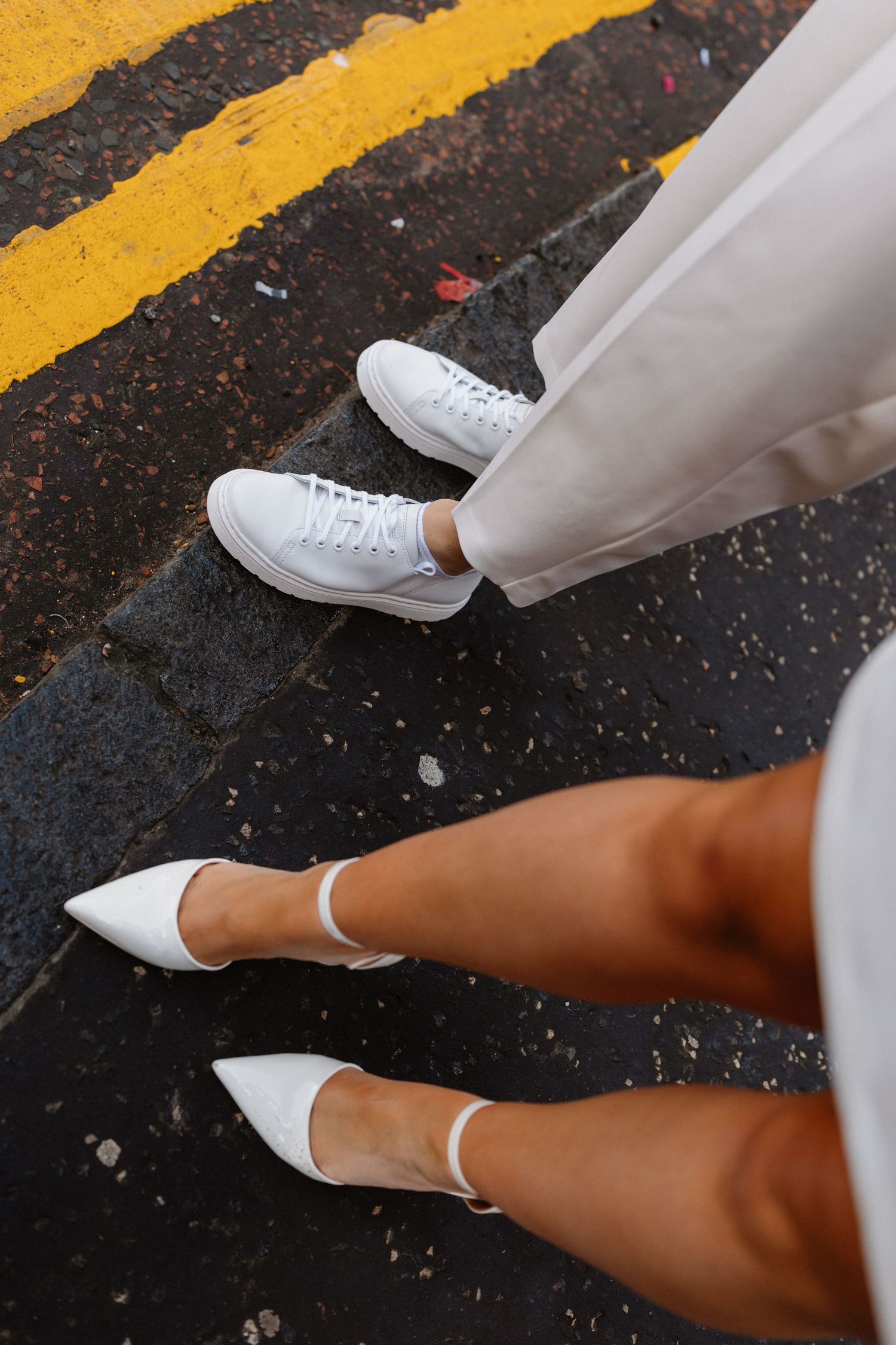 Two brides wearing white shoes, one in sneakers and one in heels, standing on a yellow-lined crosswalk.