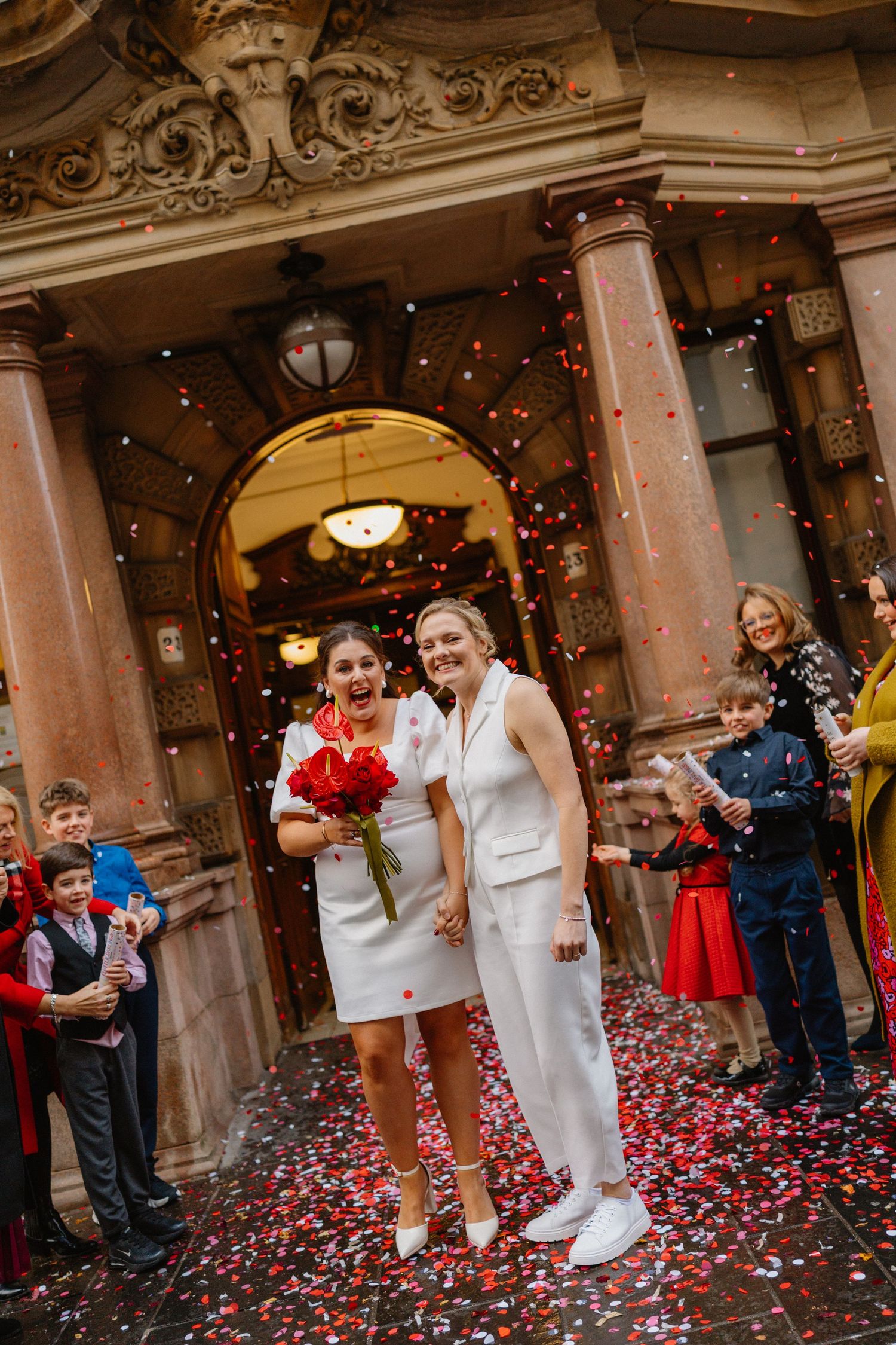 Two brides in white hold hands as colorful confetti falls outside ornate building with guests cheering.