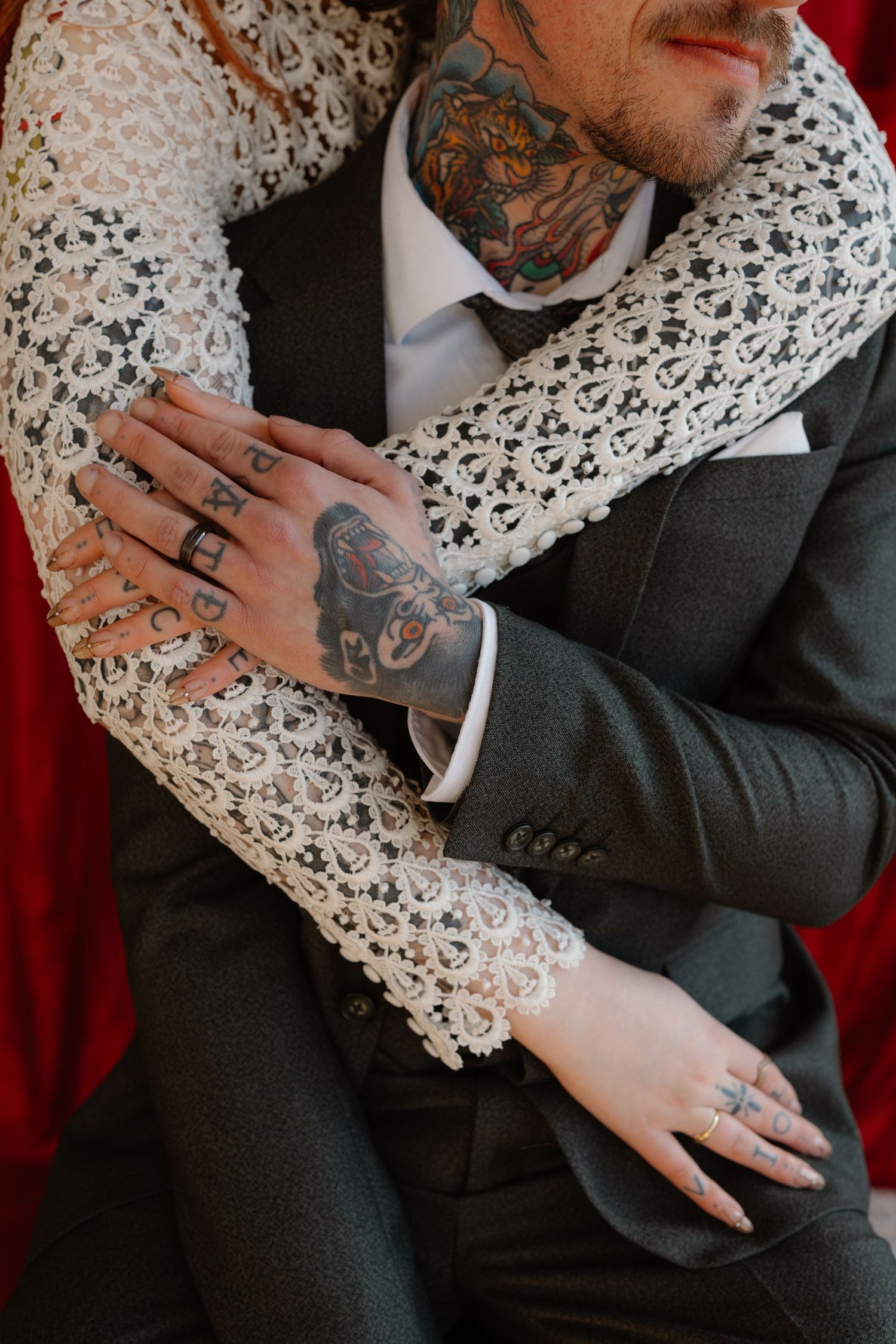 Close-up of couple embracing, showing lace wedding dress sleeve, rings, and colorful tattoos on mans hand and neck.