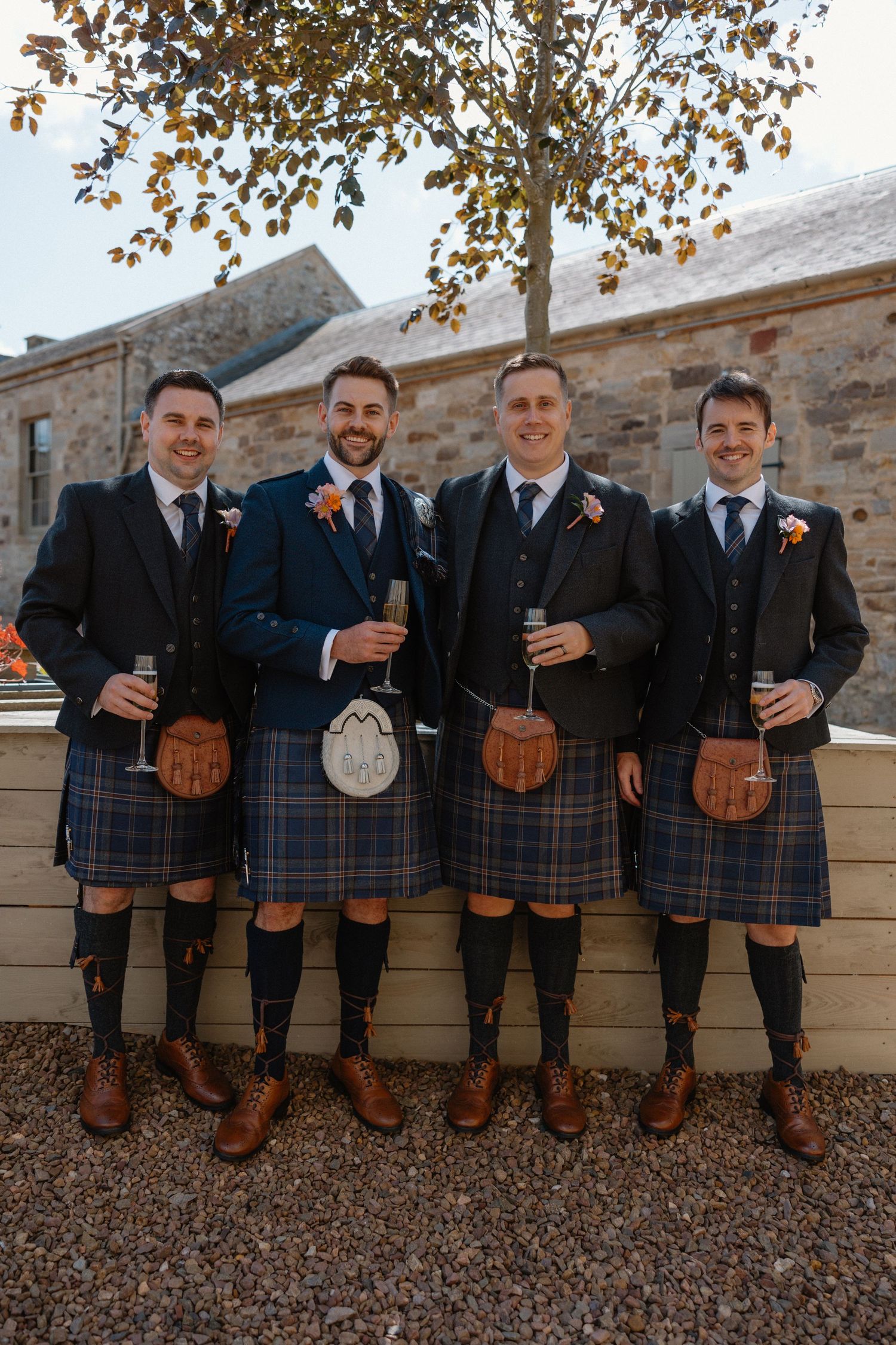 Four groomsmen in traditional Scottish kilts and dark jackets holding champagne glasses outdoors at a wedding.