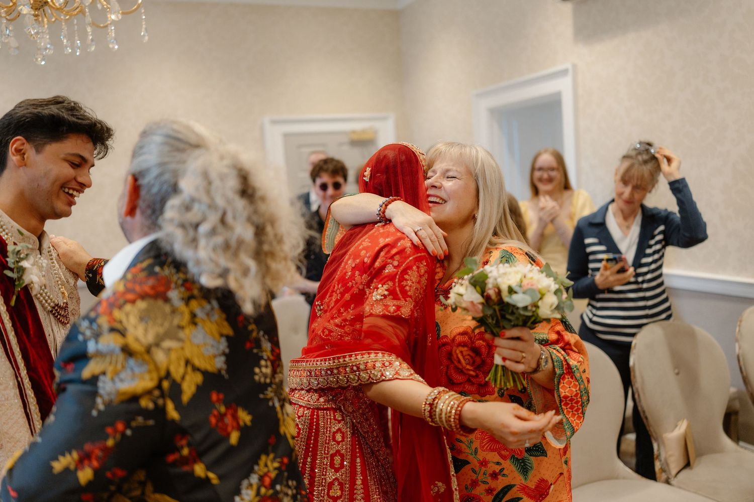 Joyful wedding guests embracing and celebrating indoors, with bride in red traditional attire holding a bouquet.