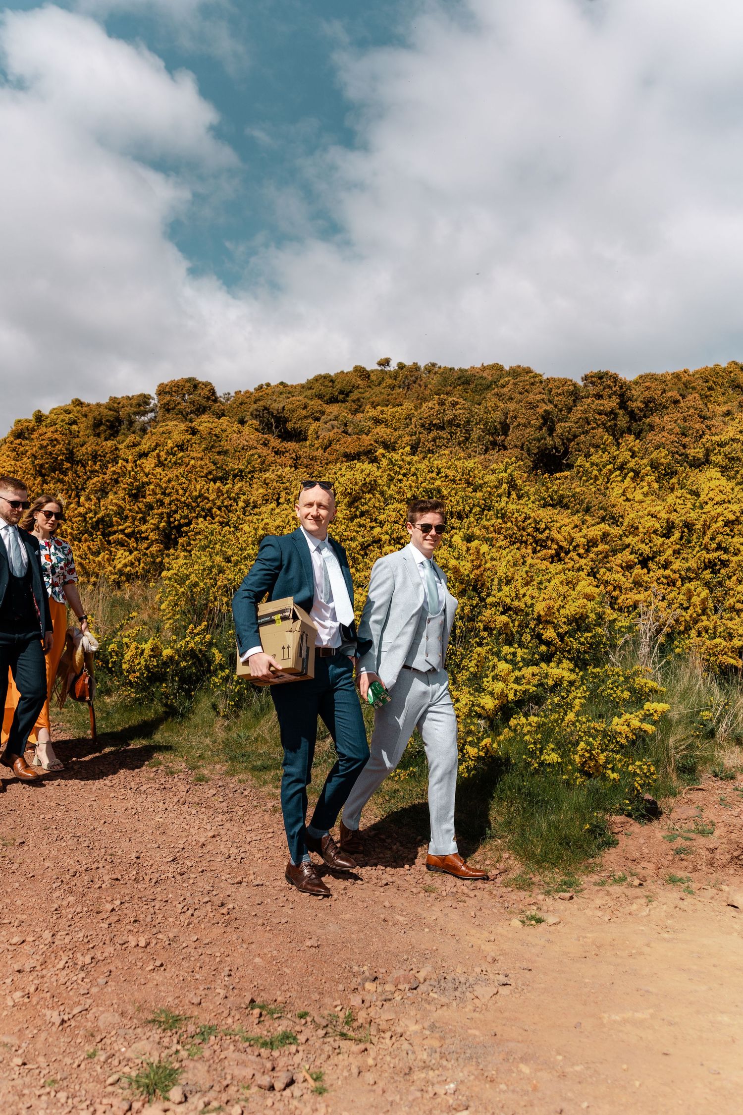 Two men in suits walk along a dirt path surrounded by yellow flowering bushes on a sunny day.